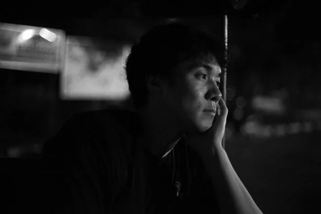 A young man sitting in a dimly lit environment, resting his face on his hand, with a contemplative expression.