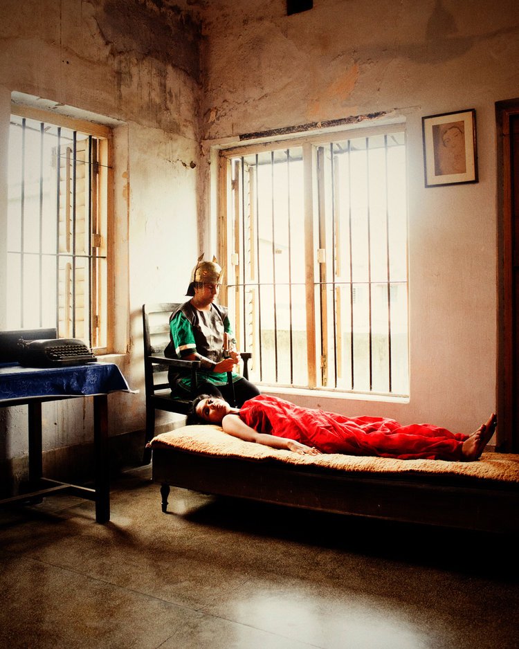A woman lies on a bed in a simple, rustic room with barred windows, while a person dressed in Roman soldier costume, including a helmet, sits nearby on a chair, holding a tablet or book. The room has minimal furnishings and a portrait hangs on the wall.