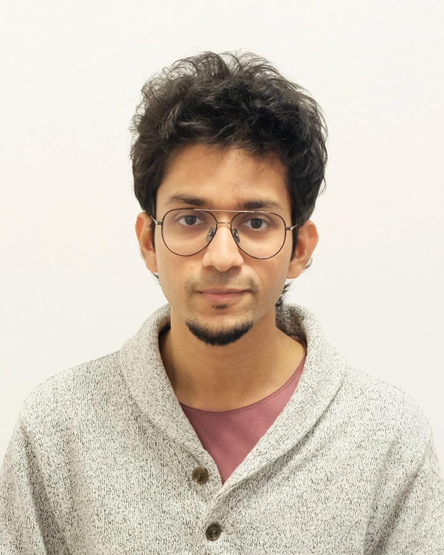 Portrait of a young man wearing glasses and a grey cardigan over a maroon shirt, standing against a plain white background.