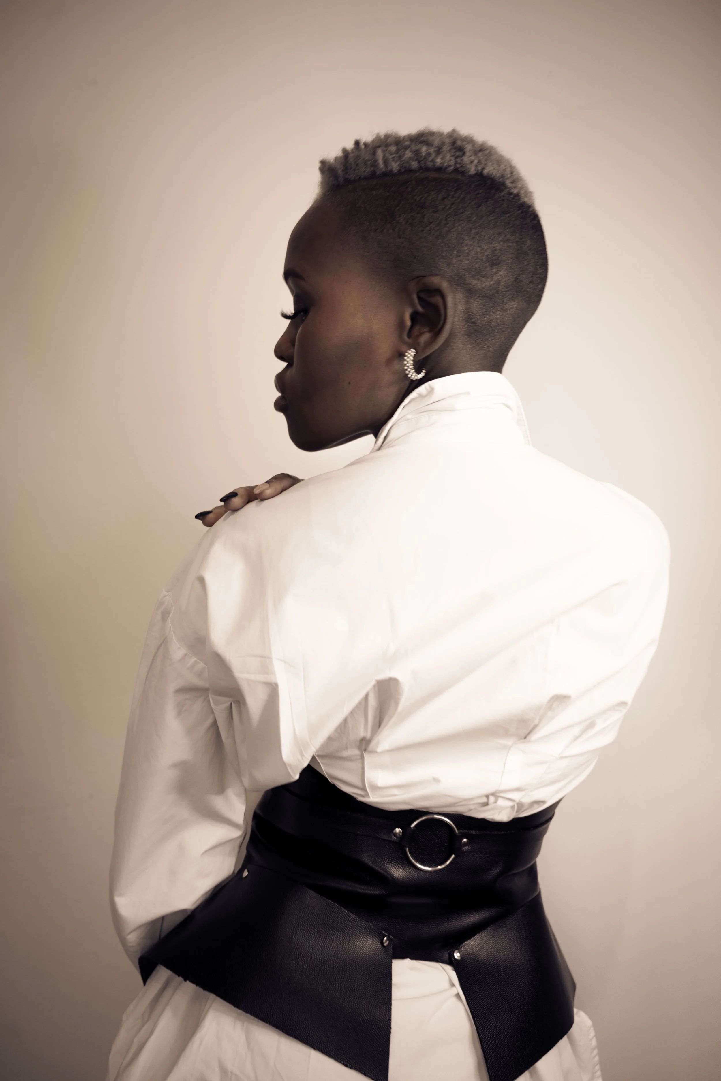 Side view of a woman with short, platinum blonde hair, wearing a white shirt and black belt with a round ring detail, standing against a plain background.
