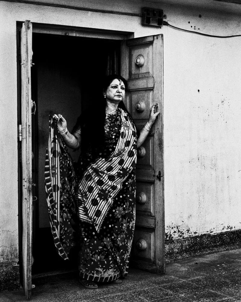 A woman in traditional Indian clothing standing in a doorway, holding the door open with one hand and a piece of fabric in the other, looking to the side.