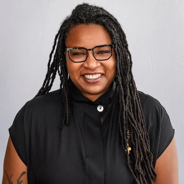 A woman with long, black dreadlocks, wearing glasses and a black button-up shirt, smiling at the camera against a plain, light-colored background.