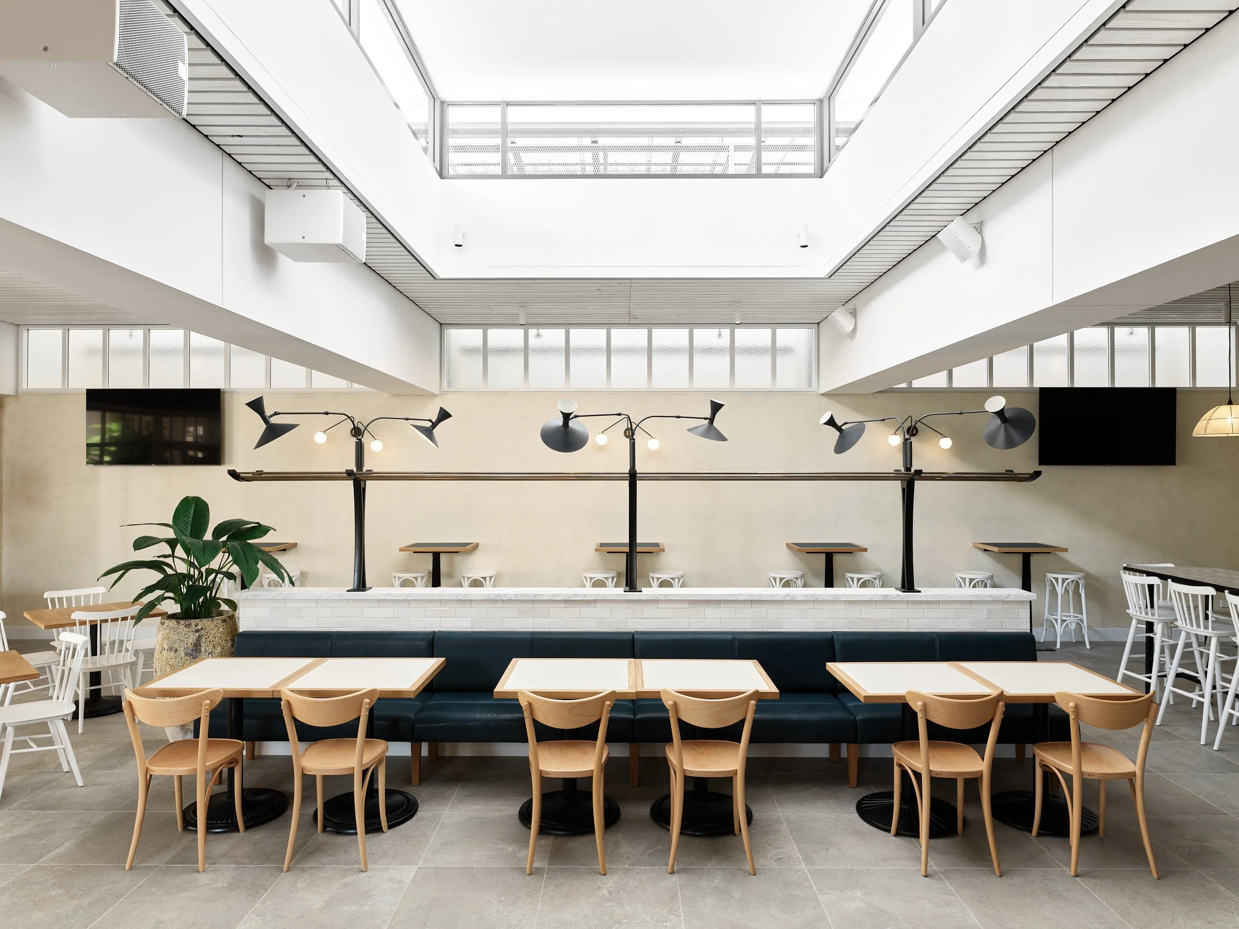 Modern cafe interior with wooden tables and chairs, black wall lamps, large windowed ceiling, potted plant, and neutral color scheme.