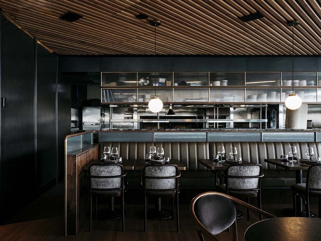 Interior of a stylish modern restaurant with dark walls, wood-paneled ceiling, and a kitchen open to the dining area. There are tables set with glasses, bowls, and utensils, and cushioned chairs around the tables.