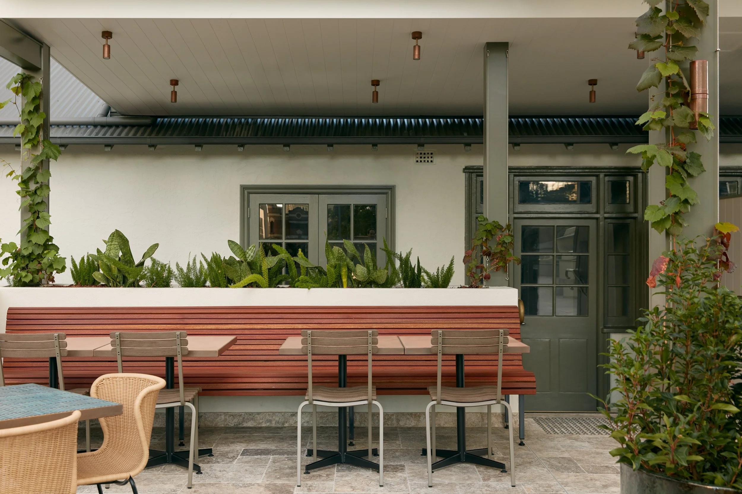 Outdoor patio area with a wooden picnic table and wicker chairs, surrounded by green plants and vines, with a building featuring a ceiling, windows, and a door in the background.