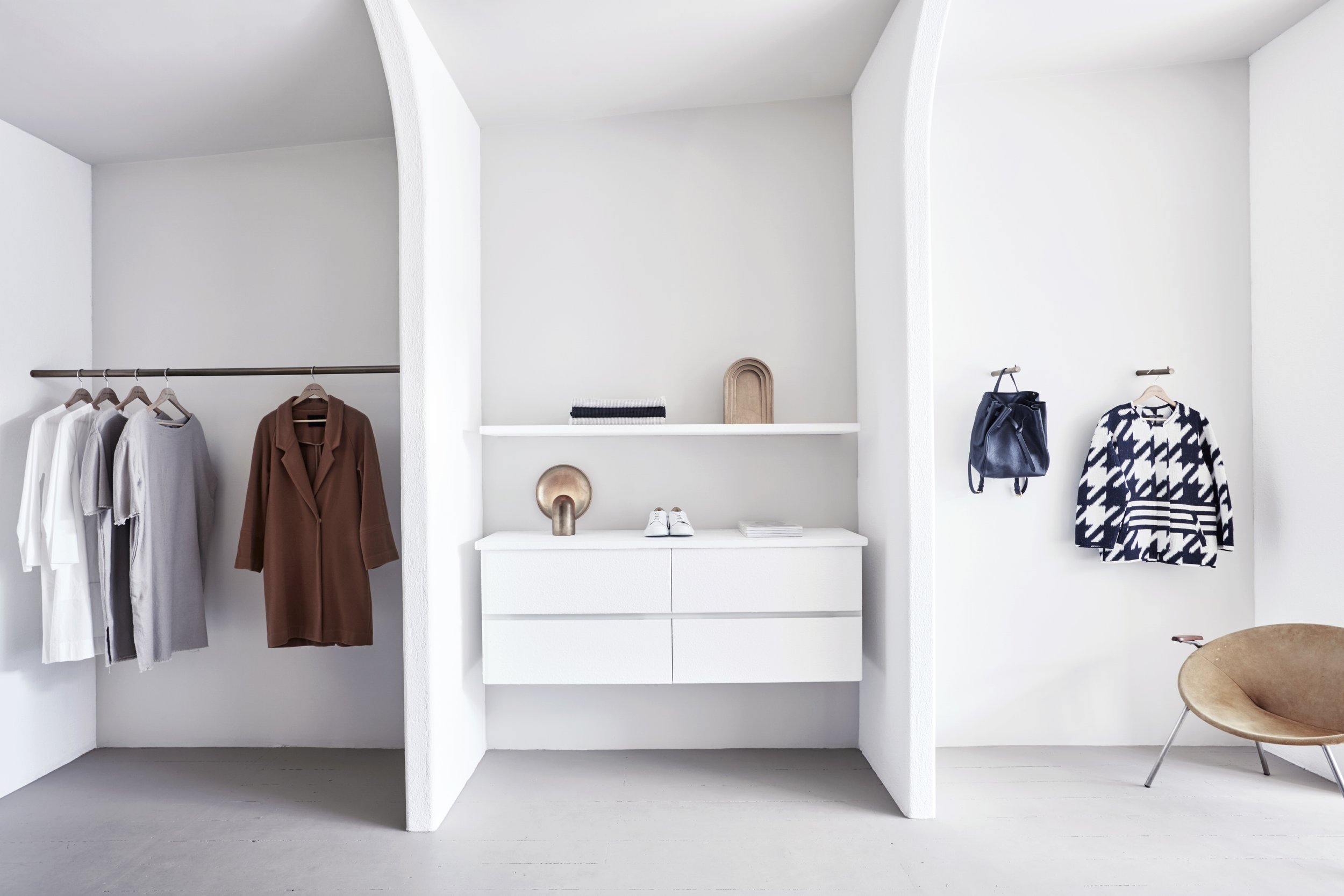 A modern, minimalistic walk-in closet with white walls and built-in storage. Clothing items hang on rods, including white, gray, brown, and navy and white patterned shirts and jackets. A small white dresser with shoes and decorative items on top is i