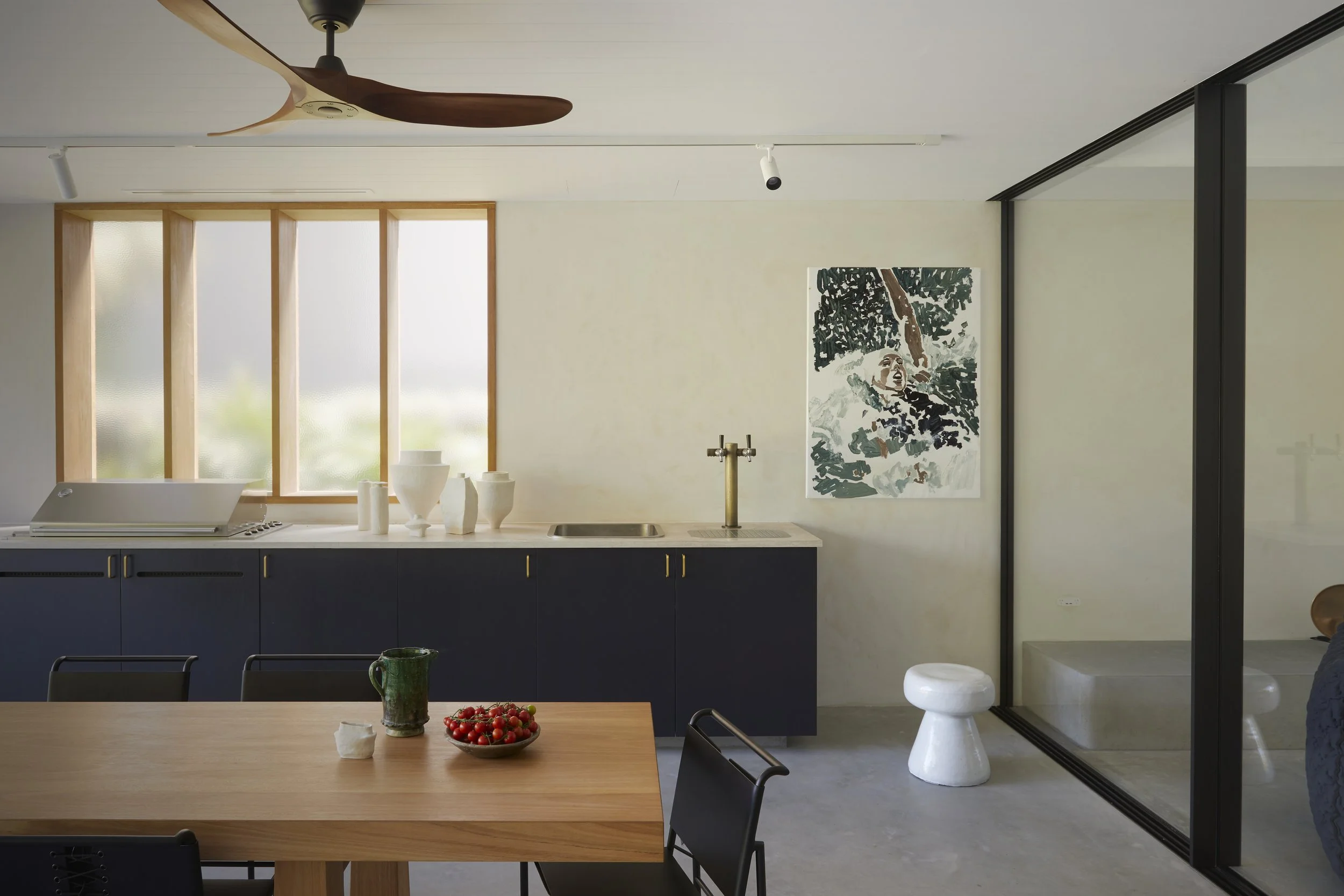 A modern kitchen and dining area with a wooden table, black chairs, and a bowl of cherry tomatoes on the table. The kitchen has dark cabinets, a white countertop, and a white backsplash. There are three large vertical windows with frosted glass and w