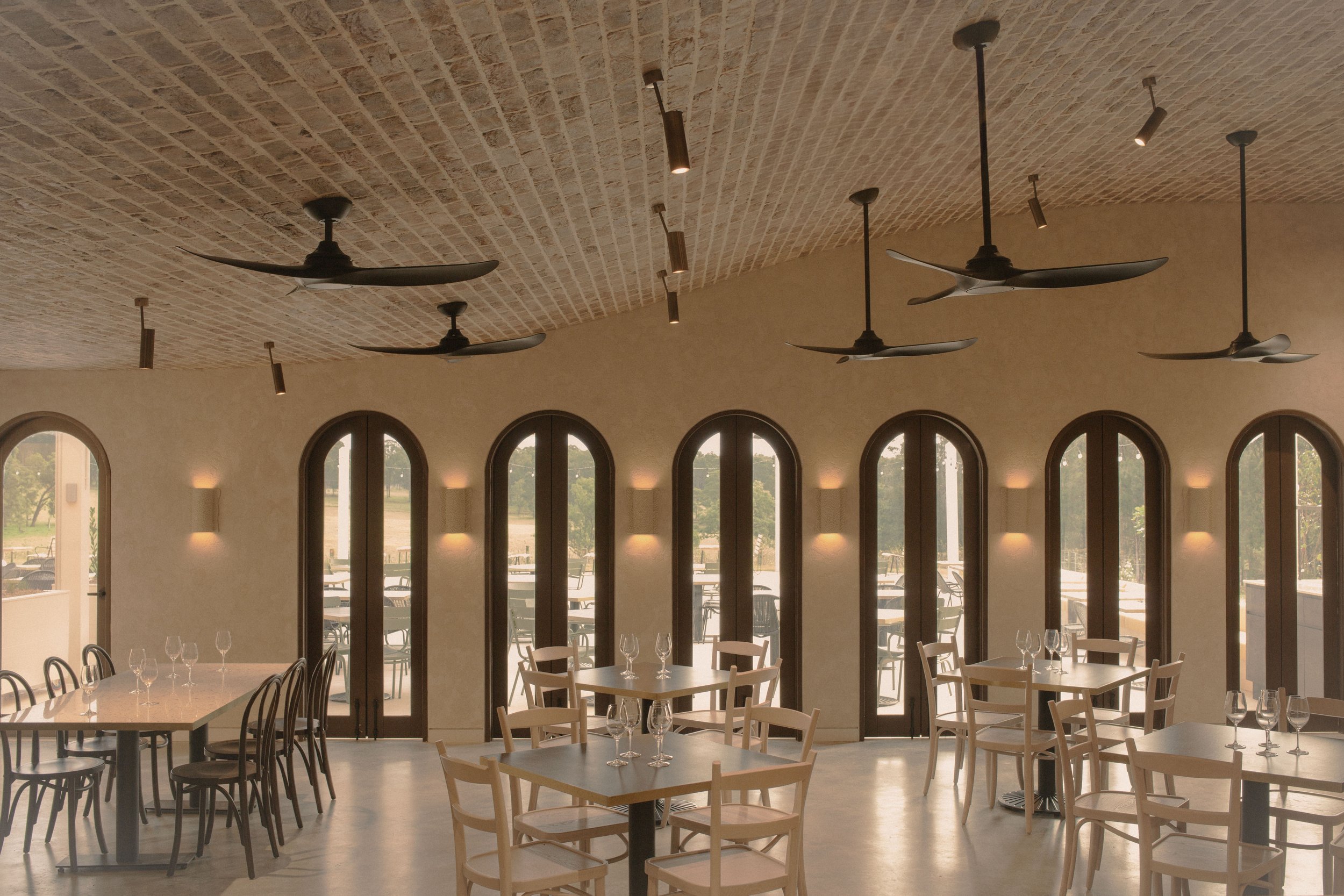Interior of a restaurant or dining area with round tables, chairs, large arched windows, ceiling fans, and hanging lights, with an outdoor patio visible through the windows.