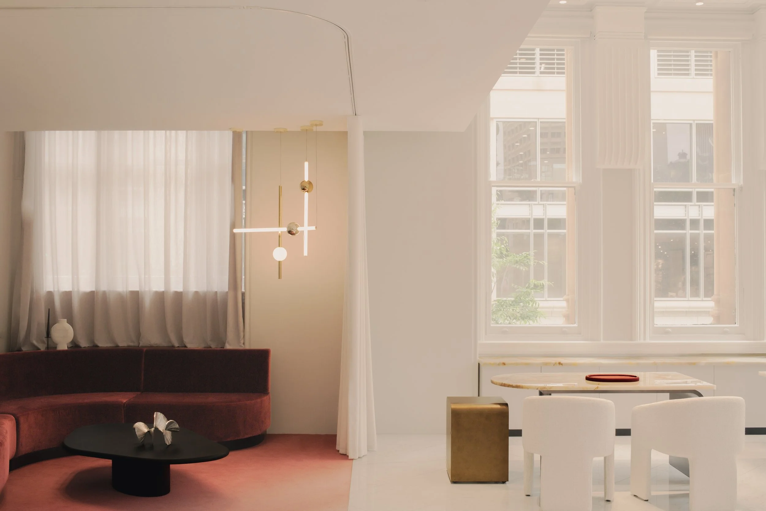 An interior space with large windows, white walls, a hanging modern light fixture, a maroon sofa, black coffee table with decorative items, and a white marble table with chairs.