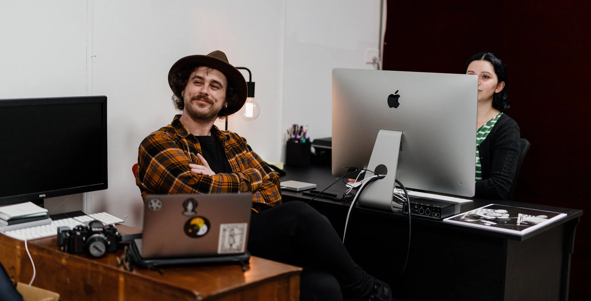 A man with curly hair, wearing a hat and plaid shirt, sitting with arms crossed at a desk in an private office, smiling, with a woman working at a large iMac computer on the other side of the room at Nauti Studios Blue Mountains