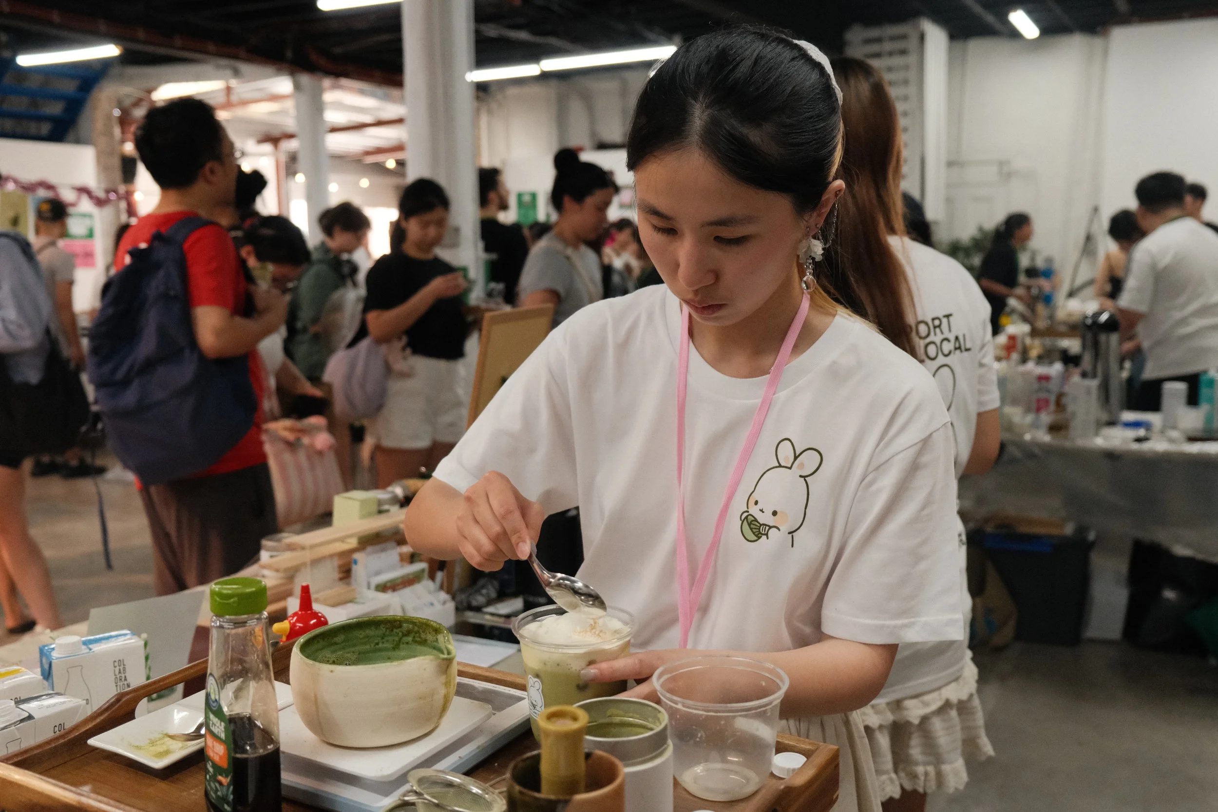 Young woman in white T-shirt preparing matcha tea at booth with green tea, bowls, and utensils, surrounded by other people at market event.