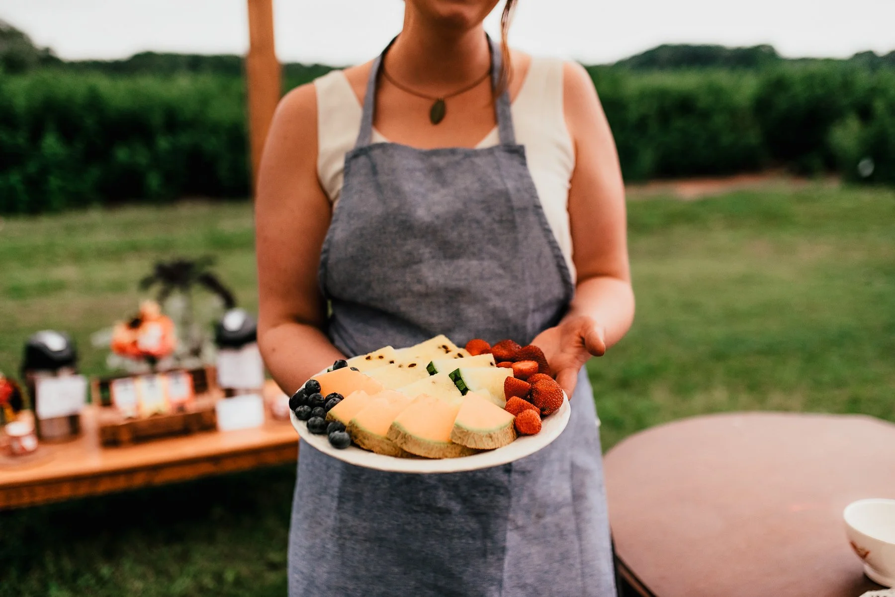 Woman wearing a blue apron holding a plate of sliced fruit, with coffee set up in the background