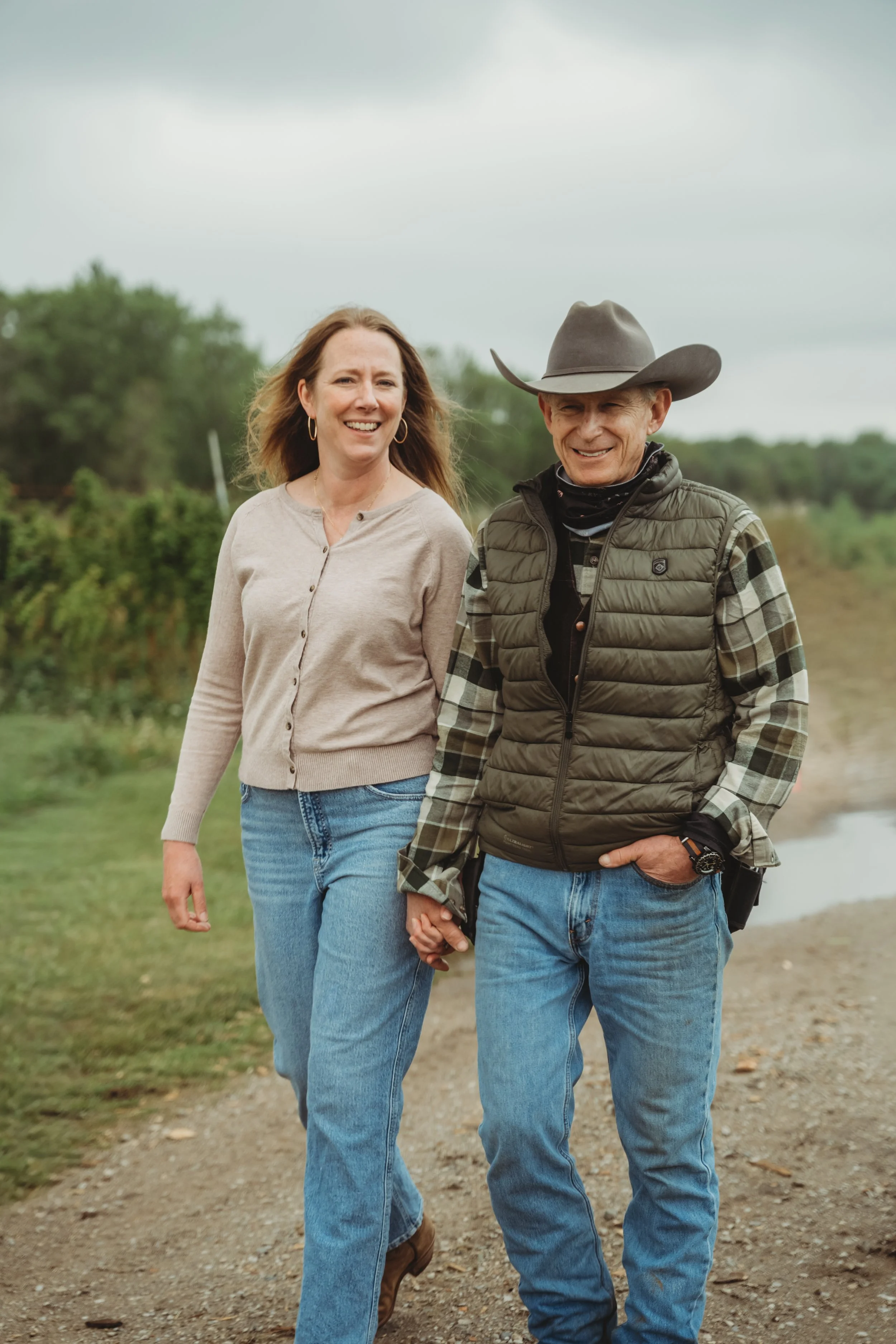 A woman with blonde hair, wearing sunglasses, a gray jacket, and a t-shirt with a graphic, standing next to a man in a white cowboy hat, plaid shirt, and brown vest. They are outdoors with a blurred background of trees and a house.