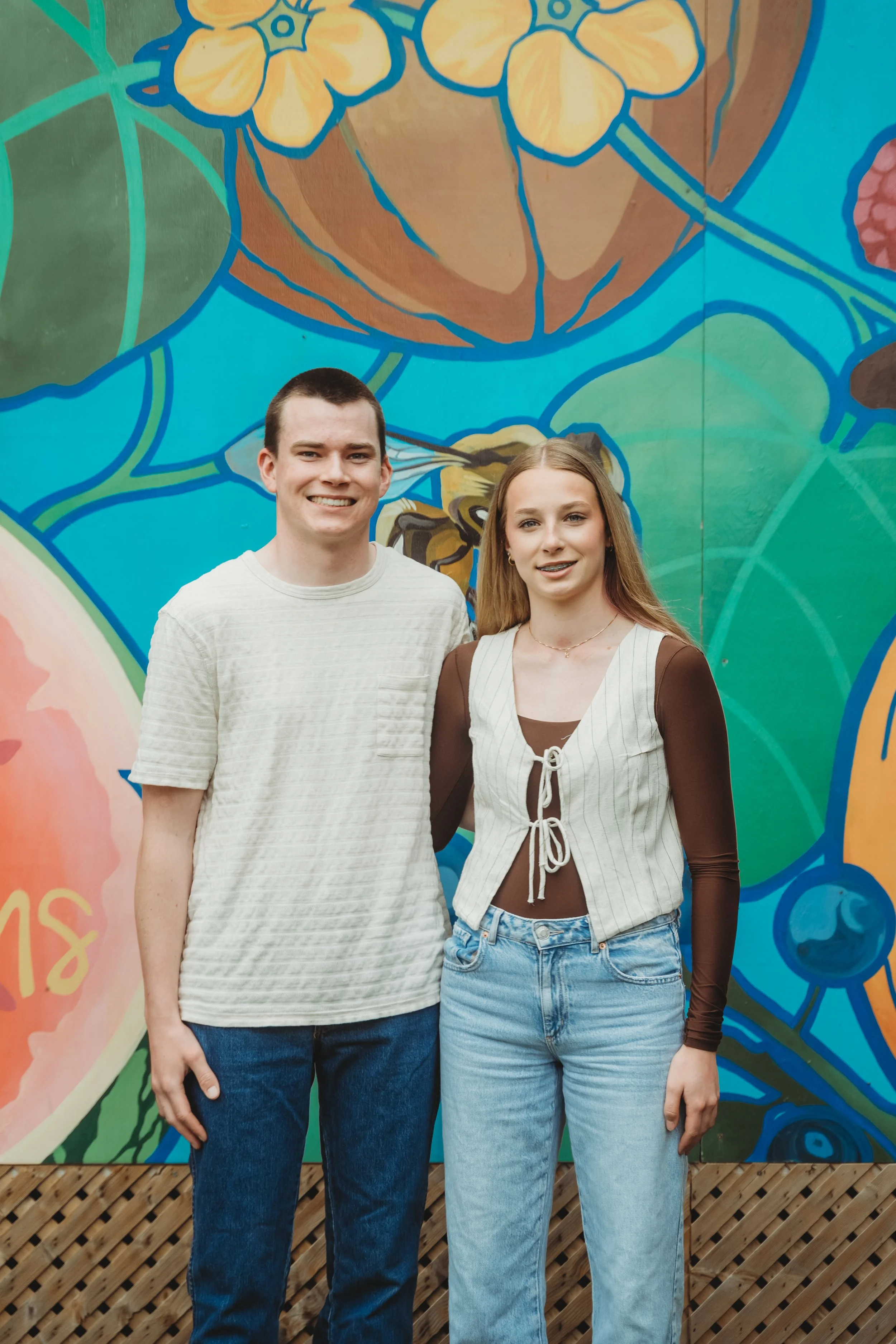 A young man and woman smiling outdoors with a cloudy sky in the background.