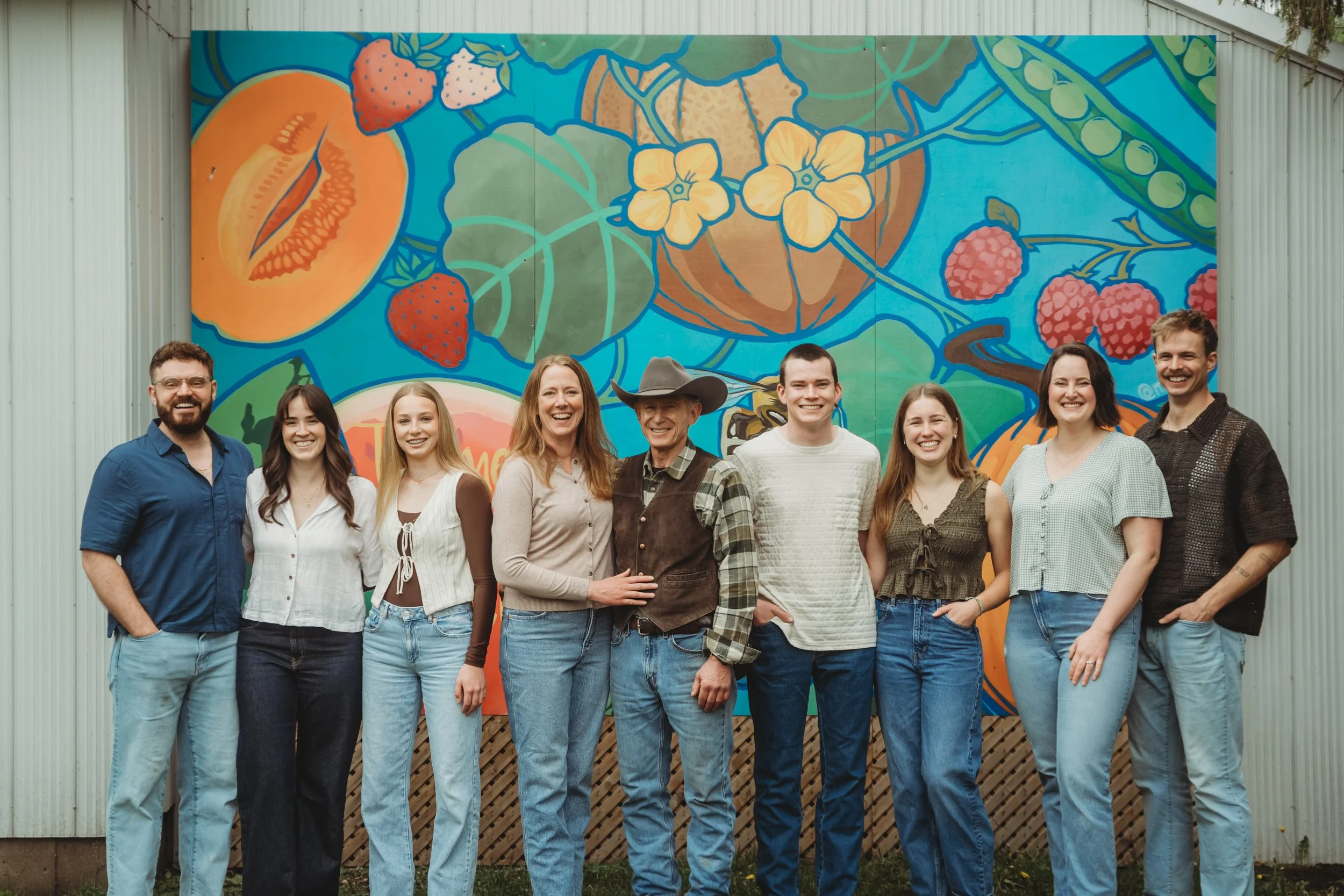 A group of 9 people smiling and standing in front of a mural showcasing fruits and vegetables