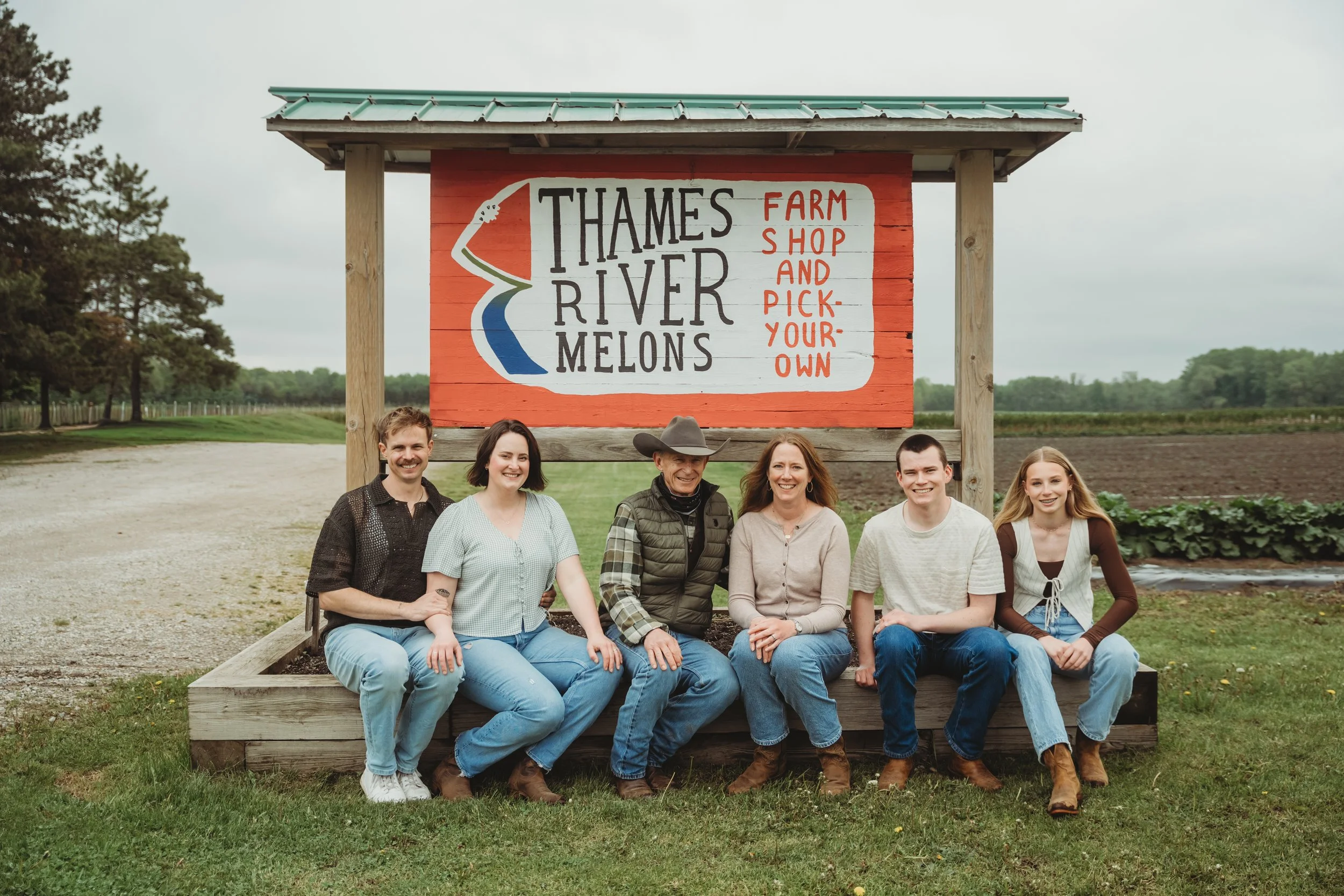 A group of 6 people sitting in front of a sign that says "Thames River Melons"