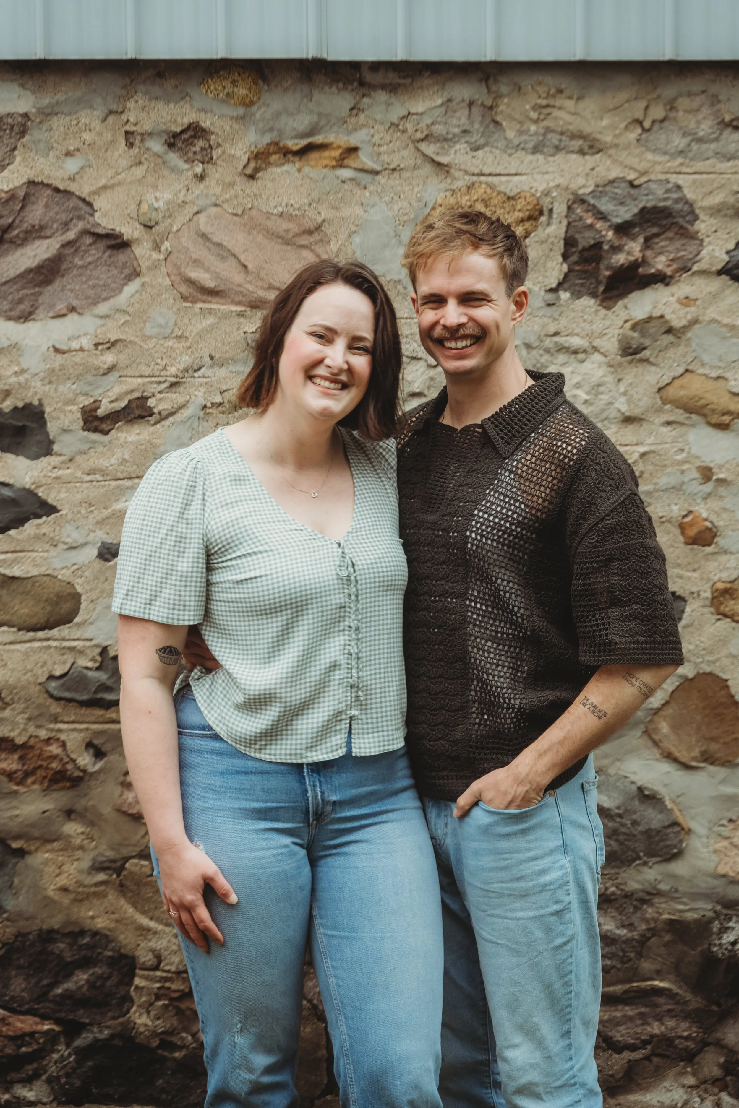 A happy couple standing close together in front of a colorful mural, smiling for the camera. The man has short brown hair and a mustache, wearing a white t-shirt. The woman has long dark hair and is wearing a plaid shirt.