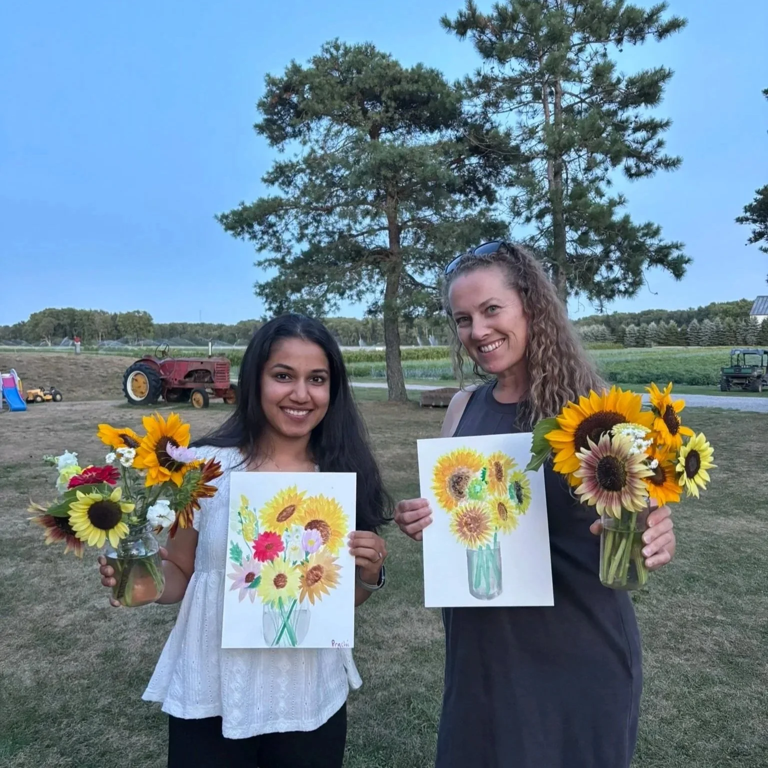 Two women smiling outdoors holding colorful sunflower bouquets and sunflower paintings on paper after their Pick 'n' Paint class at Thames River Melons in Innerkip, Ontario.