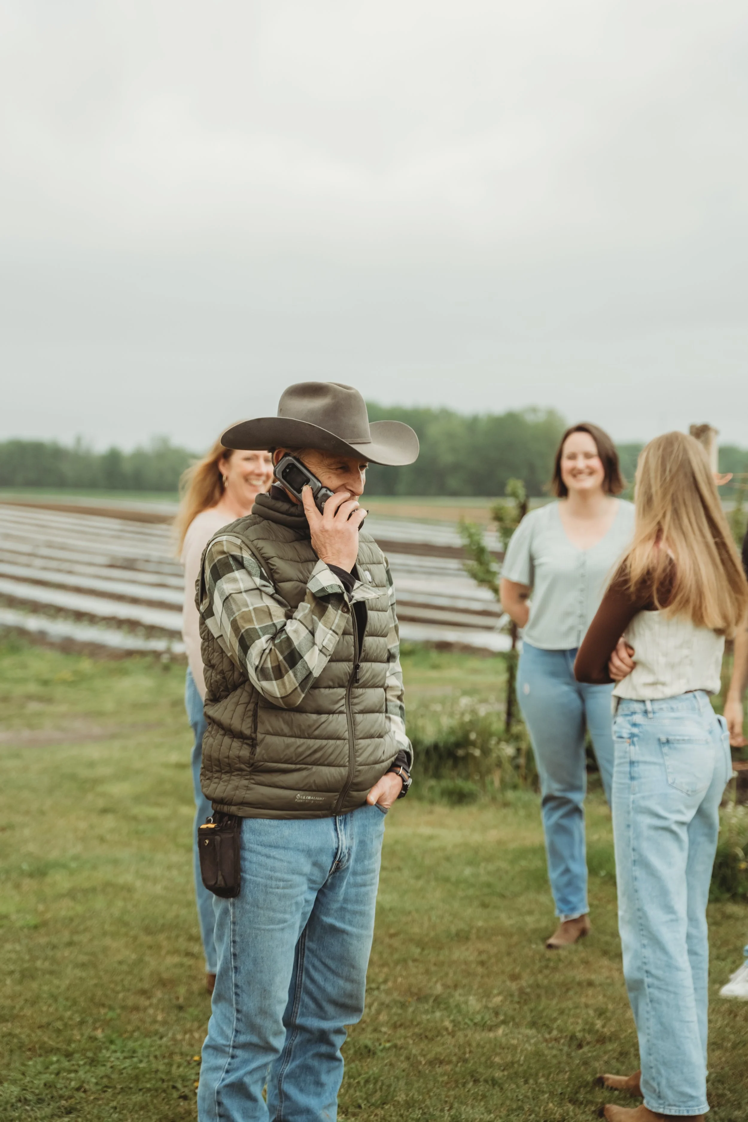 Man wearing cowboy hat, vest, and jeans talking on the phone, surrounded by 3 other people chatting in the background