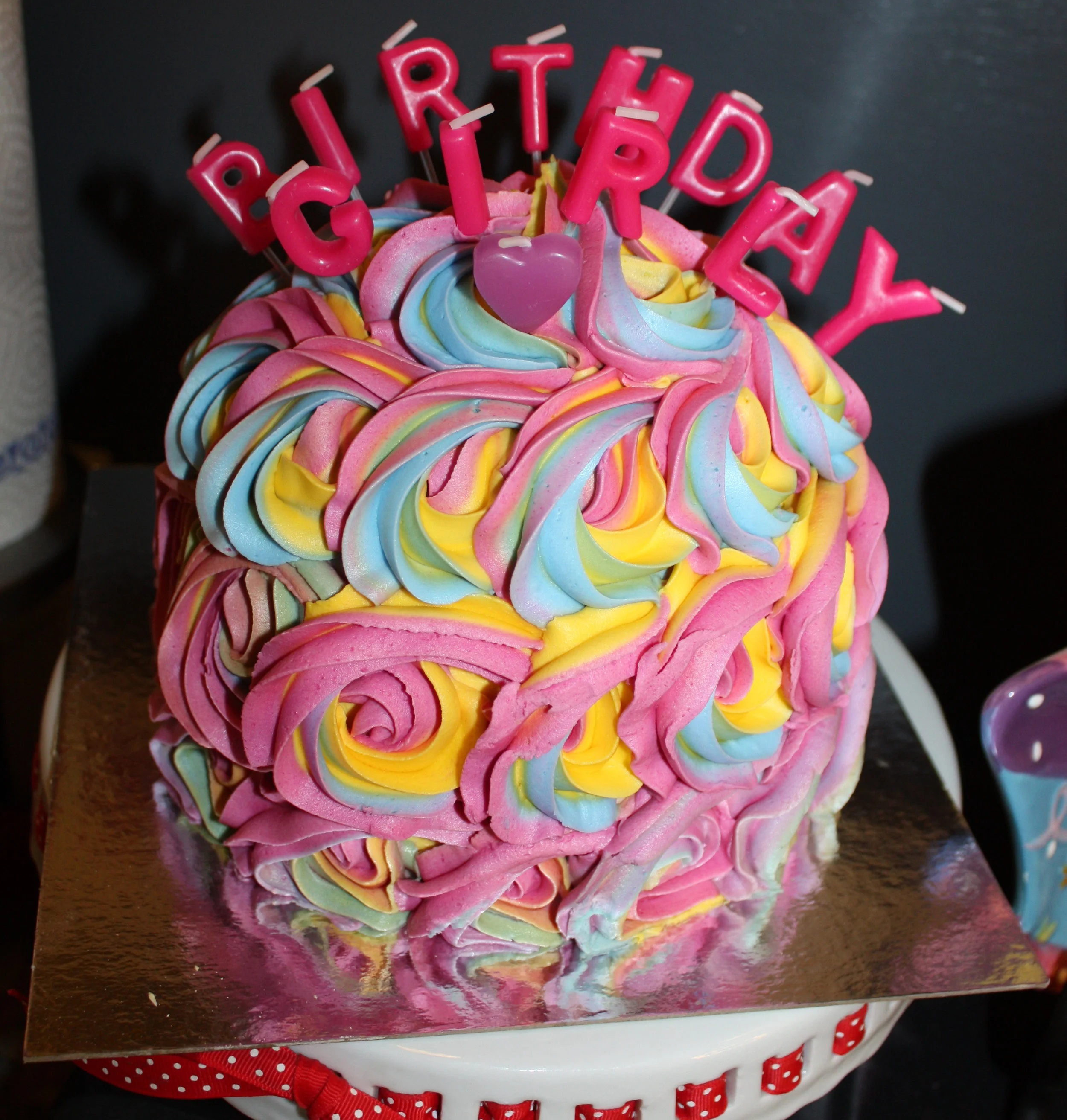 A colorful cake decorated with pastel rainbow-colored icing swirls topped with pink letters spelling 'Birthday' and a pink heart-shaped candle.