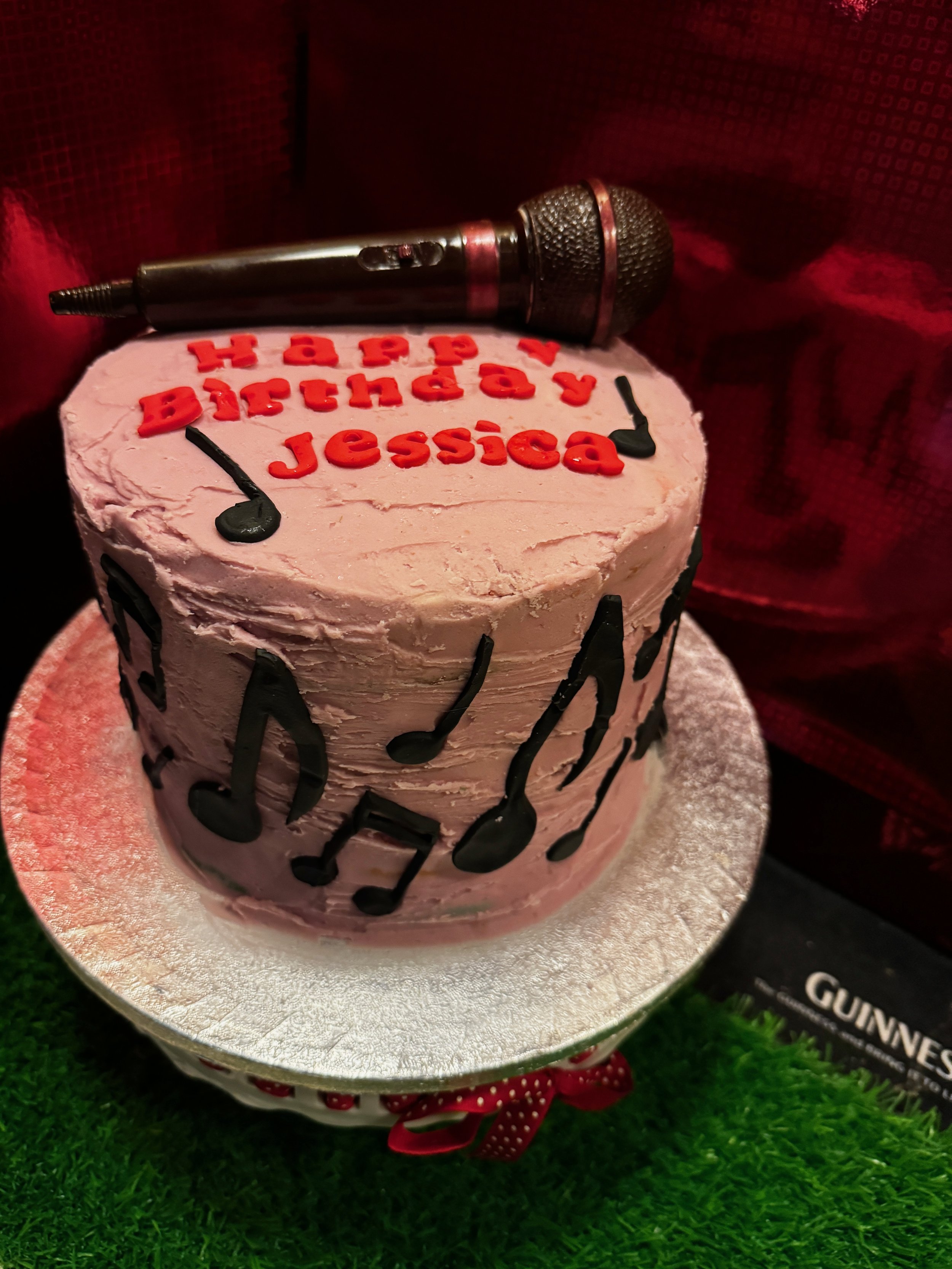 Close-up of a pink, viking-themed cake with black musical notes on it, topped with a metallic microphone decoration, on a silver cake board with green grass underneath.