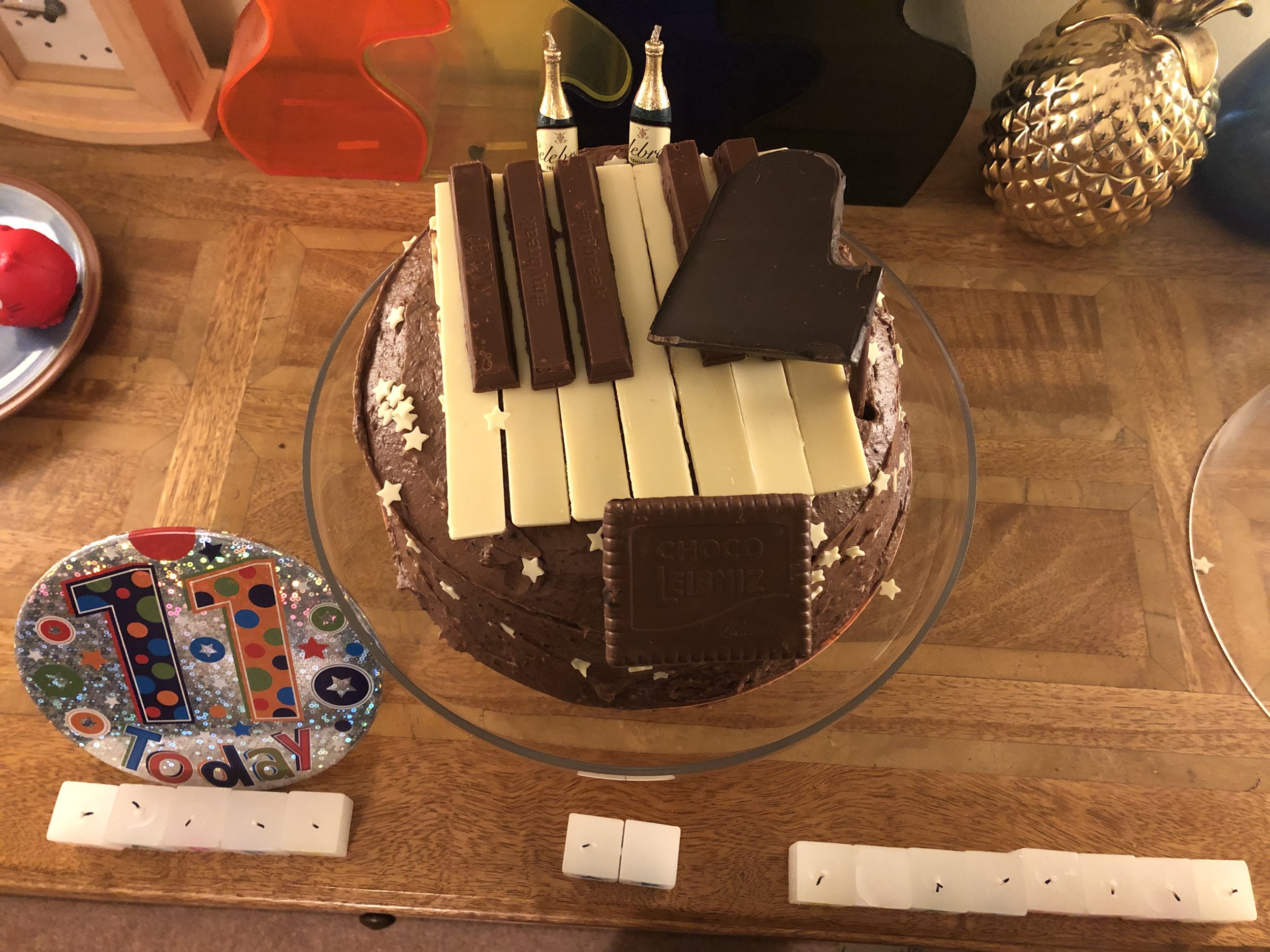 A chocolate birthday cake decorated with white and brown chocolate bars on top. A large golden pineapplike ornament to the right, a colorful 'Happy Birthday' decoration on the left, and small white blocks at the bottom.