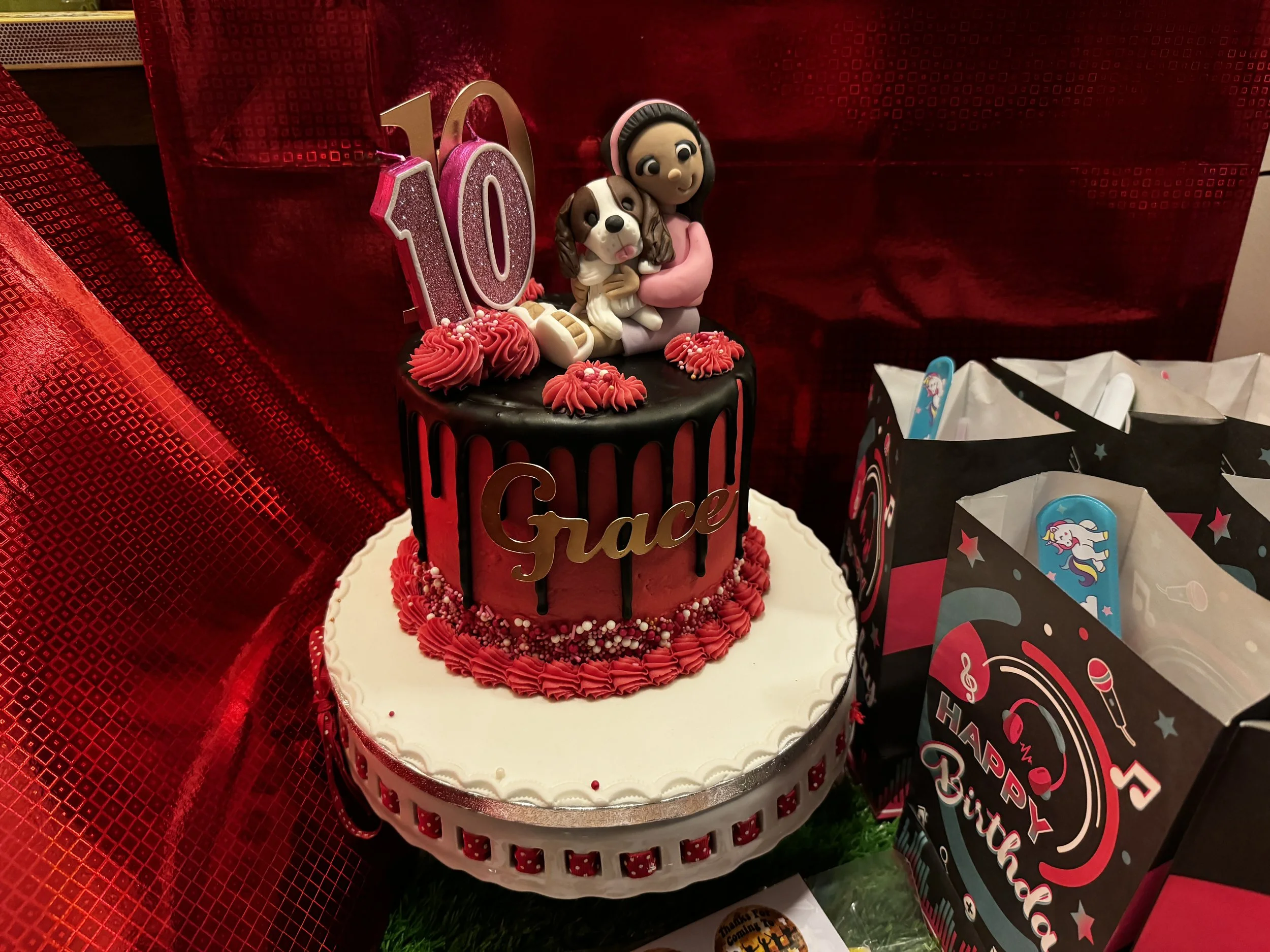 A birthday cake decorated with red and white icing, featuring a Fortnite-themed topper with a girl and a dog, and decorated with the number '10' candles, on a white and red cake stand. Red textured gift wrapping in the background.