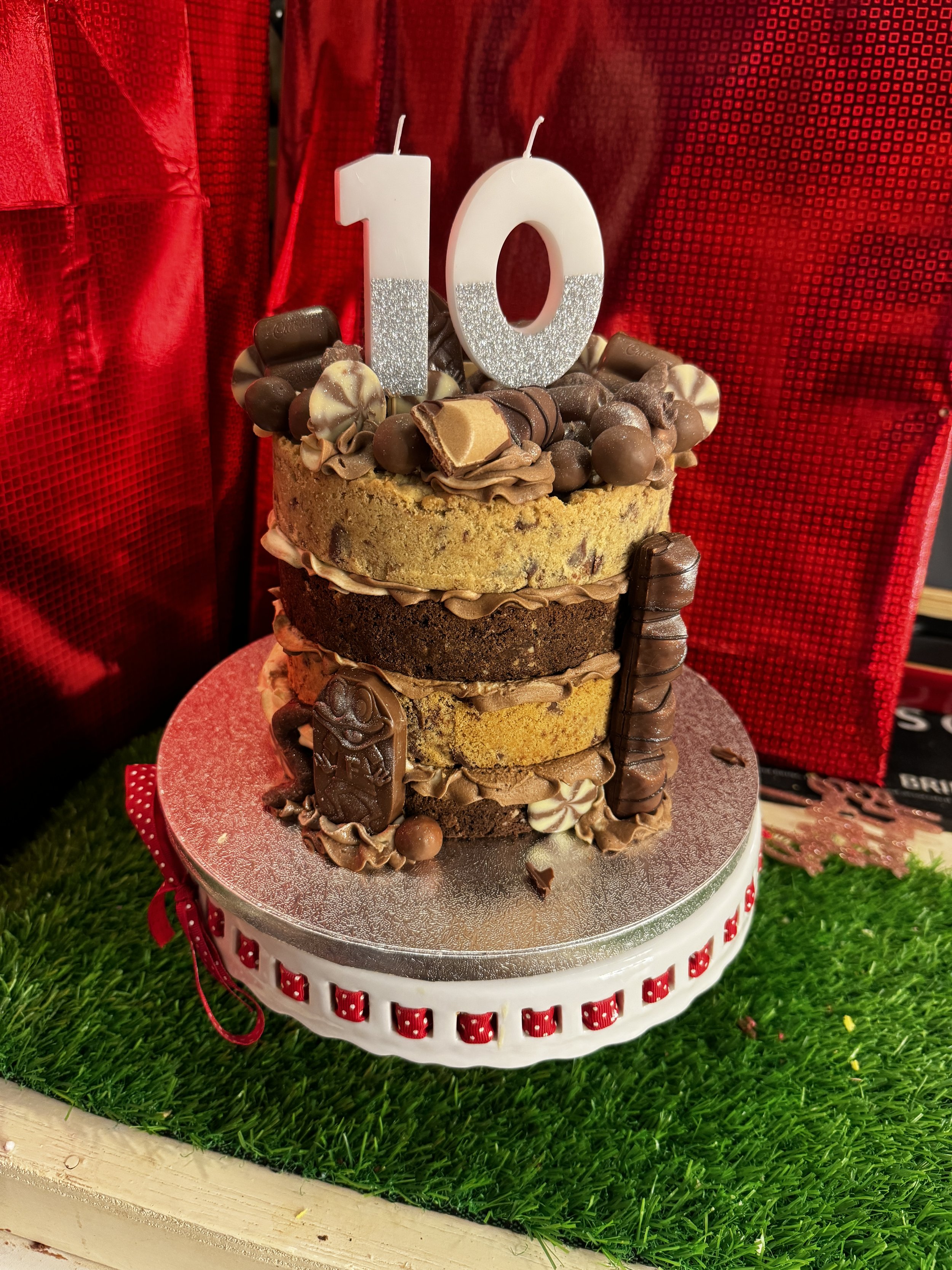A multi-layer chocolate and vanilla birthday cake decorated with various chocolates, candies, and a large white candle shaped as the number 10, on a red cake stand against a red curtain backdrop.