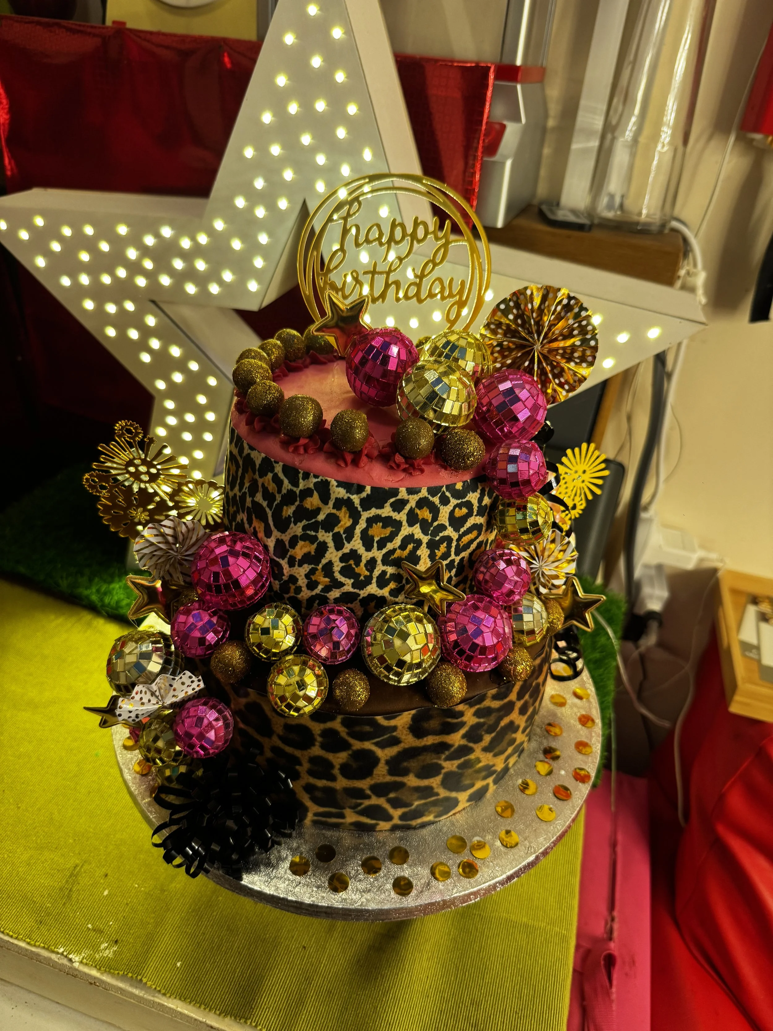 A decorated birthday cake with a leopard print pattern, topped with pink and gold disco ball ornaments, gold glittery balls, and star-shaped decorations. A gold "happy birthday" cake topper is placed on top. The cake is on a silver plate with a yellow and green surface underneath.