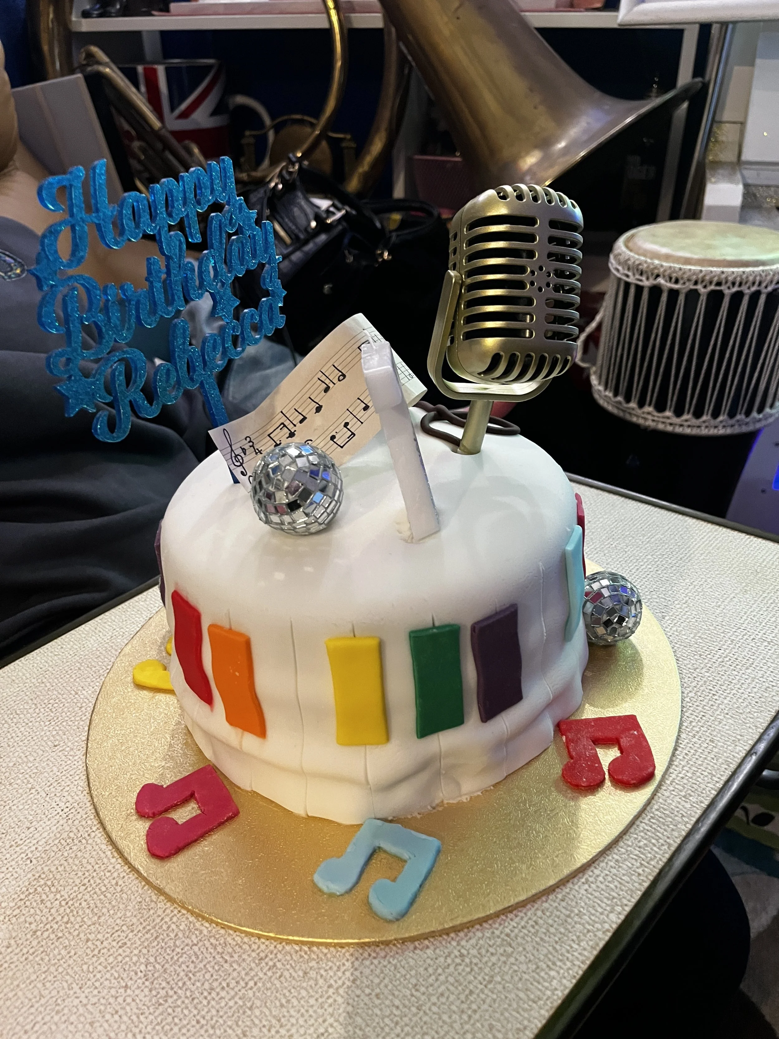 A birthday cake decorated with rainbow-colored rectangles, musical notes made of fondant on the cake and on the table, disco balls, a small portable microphone, and a blue glittery 'Happy Birthday Rebecca' topper.