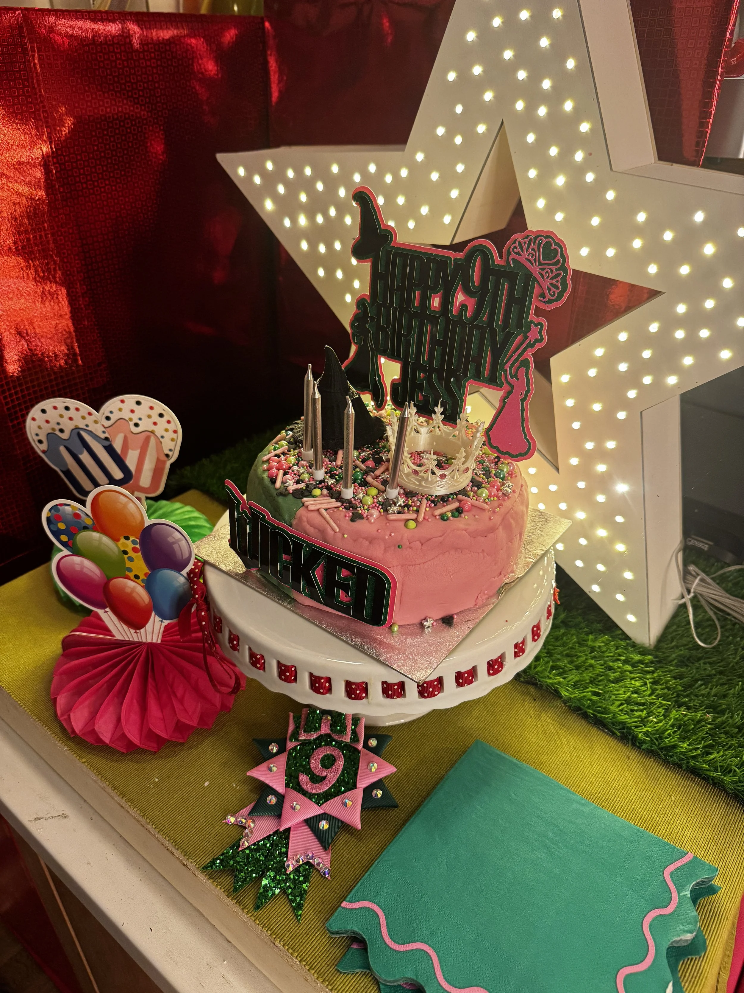 A pink birthday cake with colorful sprinkles and candles, decorated with a 'Happy Birthday' sign, surrounded by birthday-themed decorations, and illuminated star-shaped light display in the background.