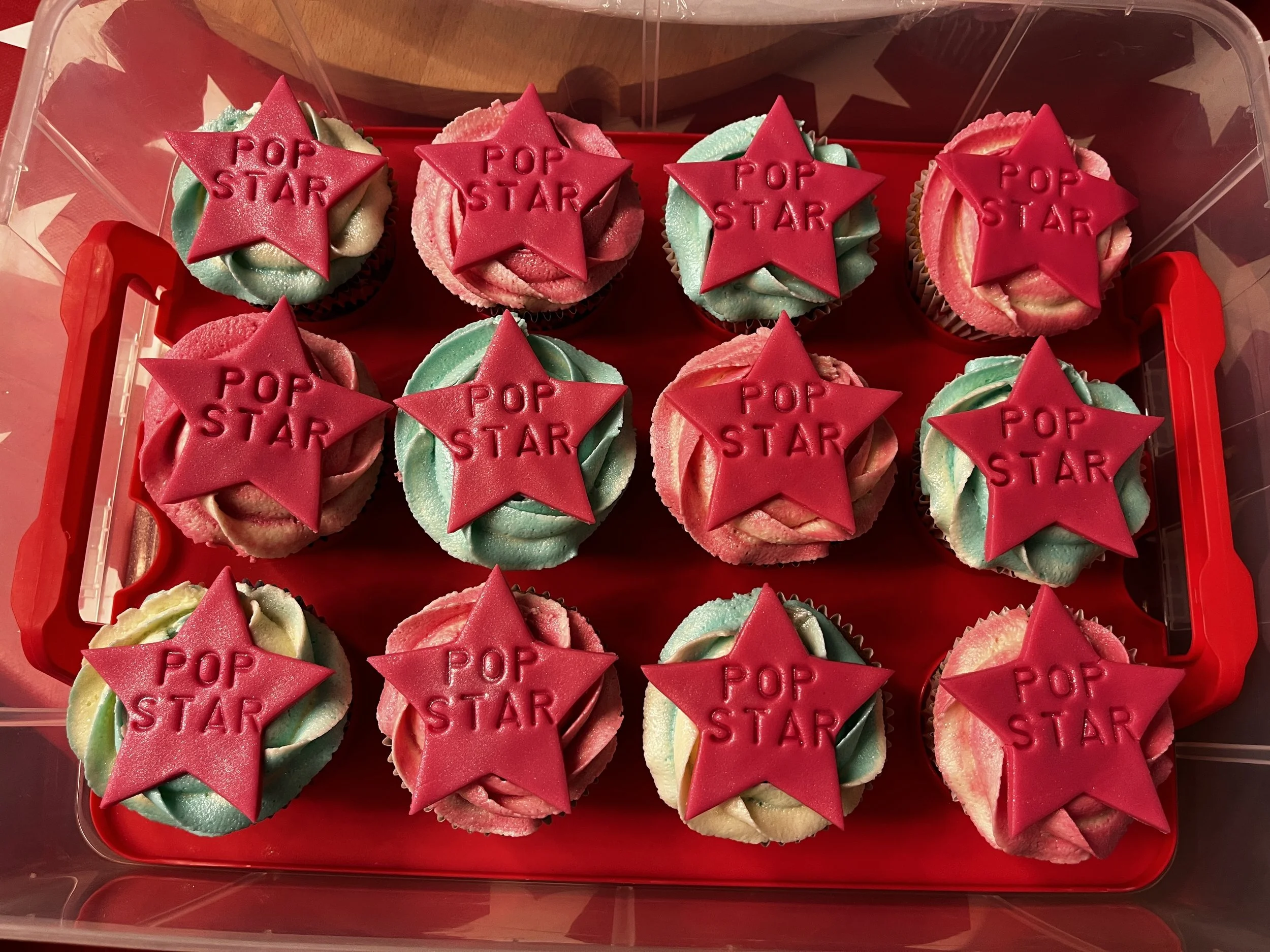 A tray of colorful cupcakes with swirled frosting in pastel shades of green, pink, and blue, topped with red star-shaped decorations that read 'POP STAR' in raised letters.