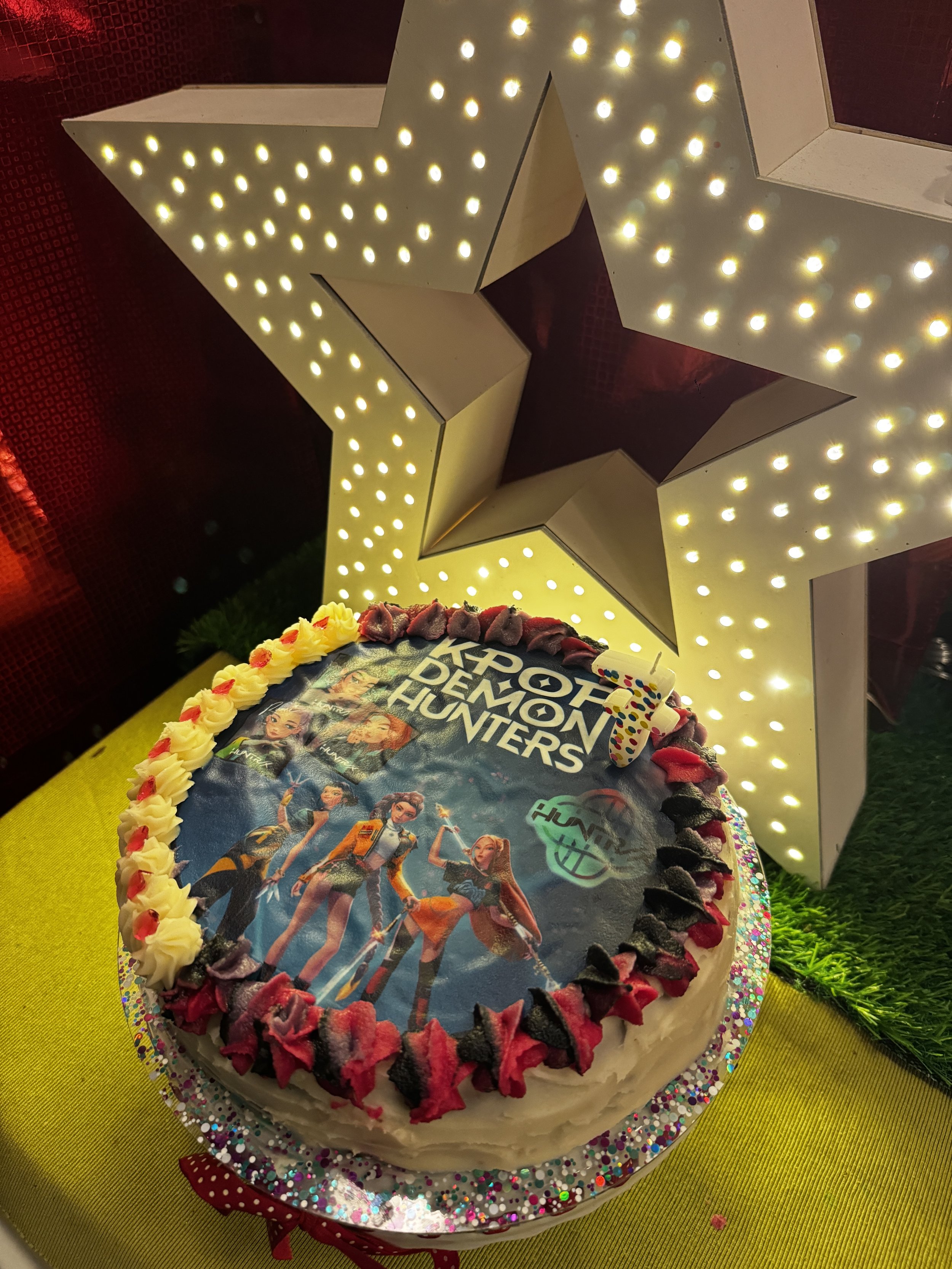 A cake decorated with a Marvel's Avengers 'X-Force Demons Hunters' themed edible topper featuring characters from the game. The cake is surrounded by colorful frosting and is placed on a yellow surface with a star-shaped decorative light in the background.