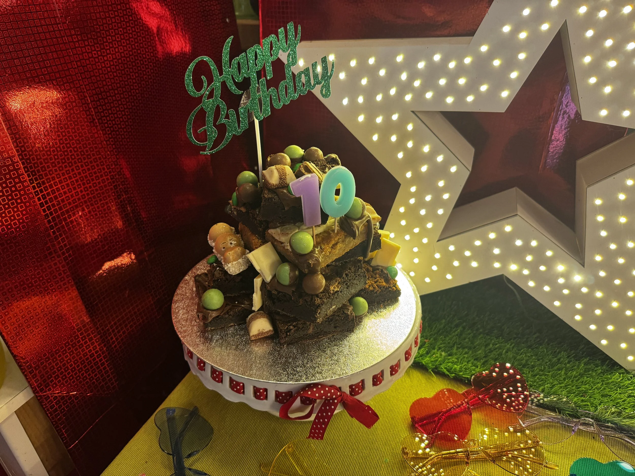 Colorful birthday cake decorated with candies and topped with a 'Happy Birthday' sign and number candles, arranged on a round silver cake stand with festive decorations around.