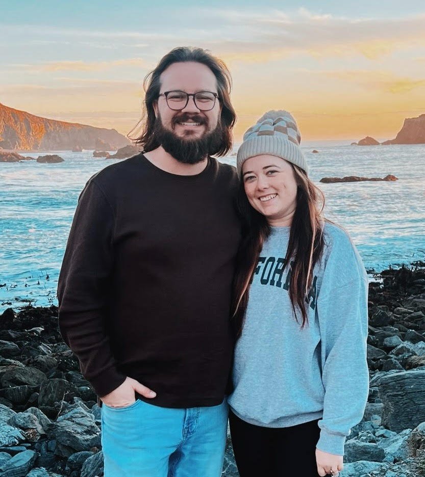 Couple Devin and Shannon at the beach, smiling at the camera at sunset