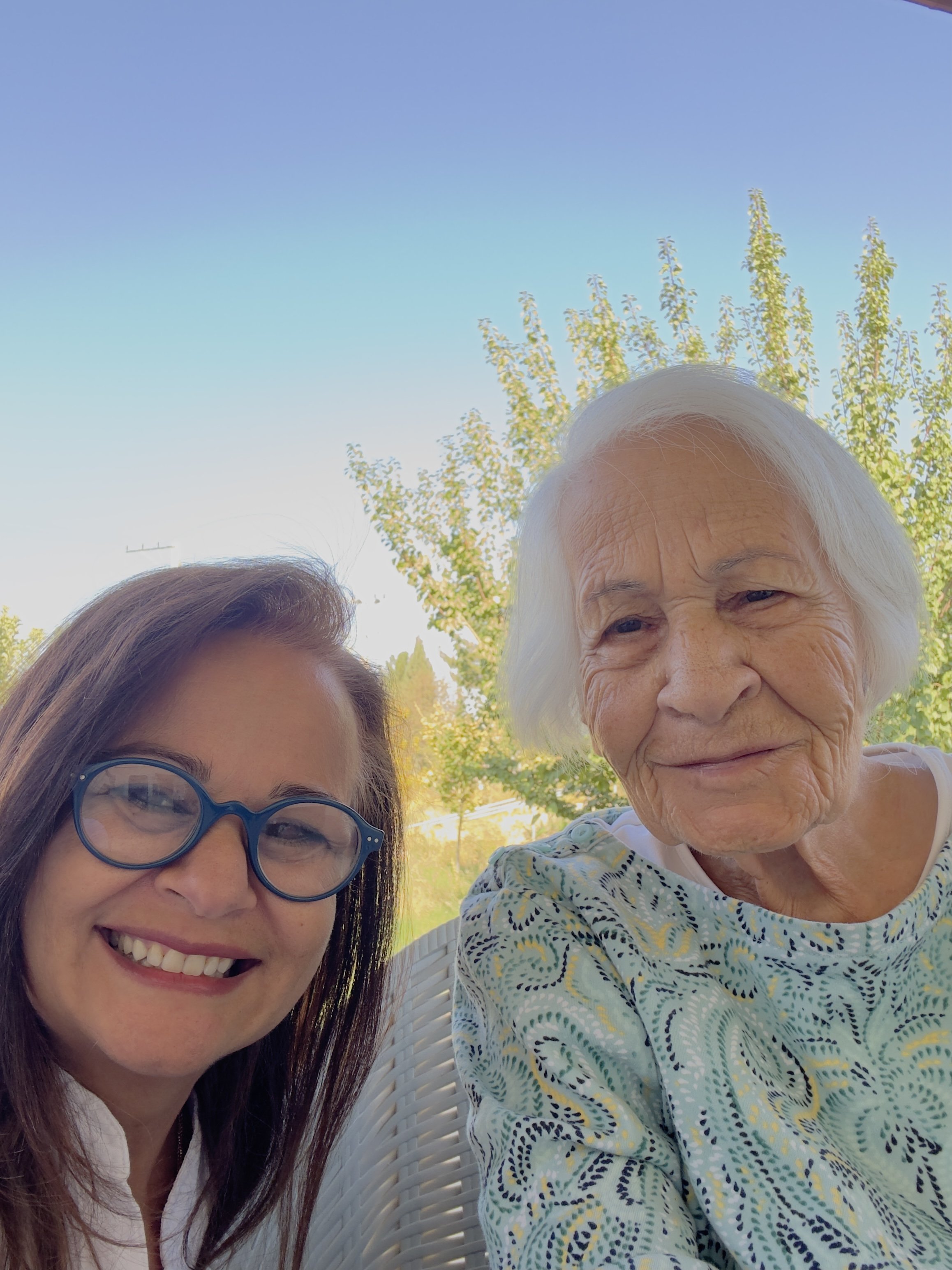 A woman with blue glasses and a smiling elderly woman with white hair sitting outdoors on a sunny day, with trees and a clear sky in the background.