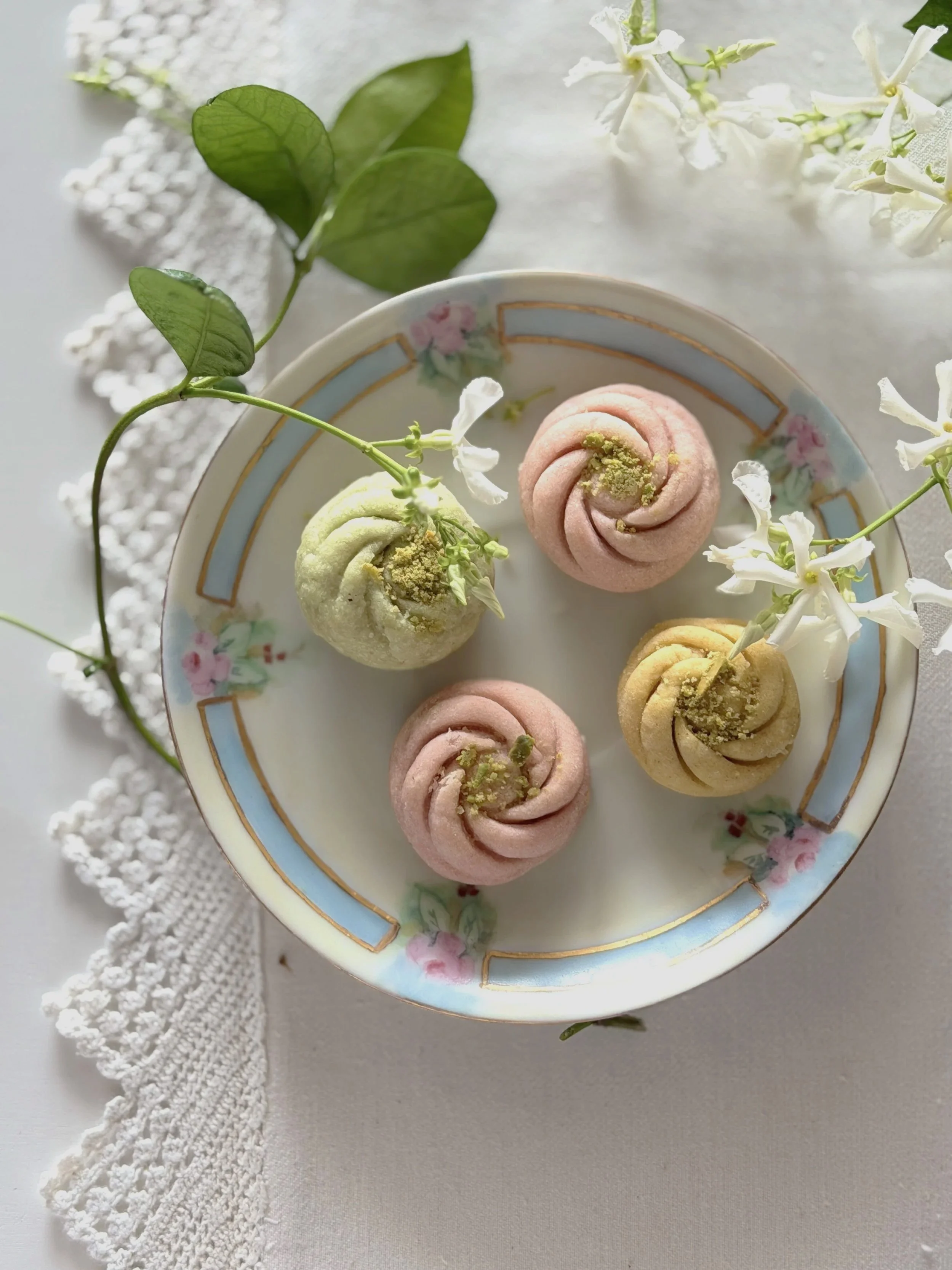 A porcelain plate with a floral design holding three colorful Japanese wagashi confections decorated with green powder and small flowers, surrounded by white flowers and green leaves on a white lace cloth.