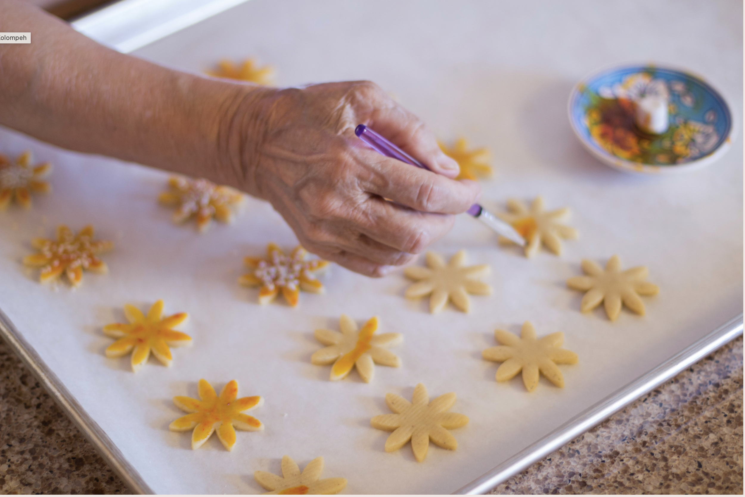 A person decorating flower-shaped cookies with orange and white icing on a baking sheet covered with parchment paper.
