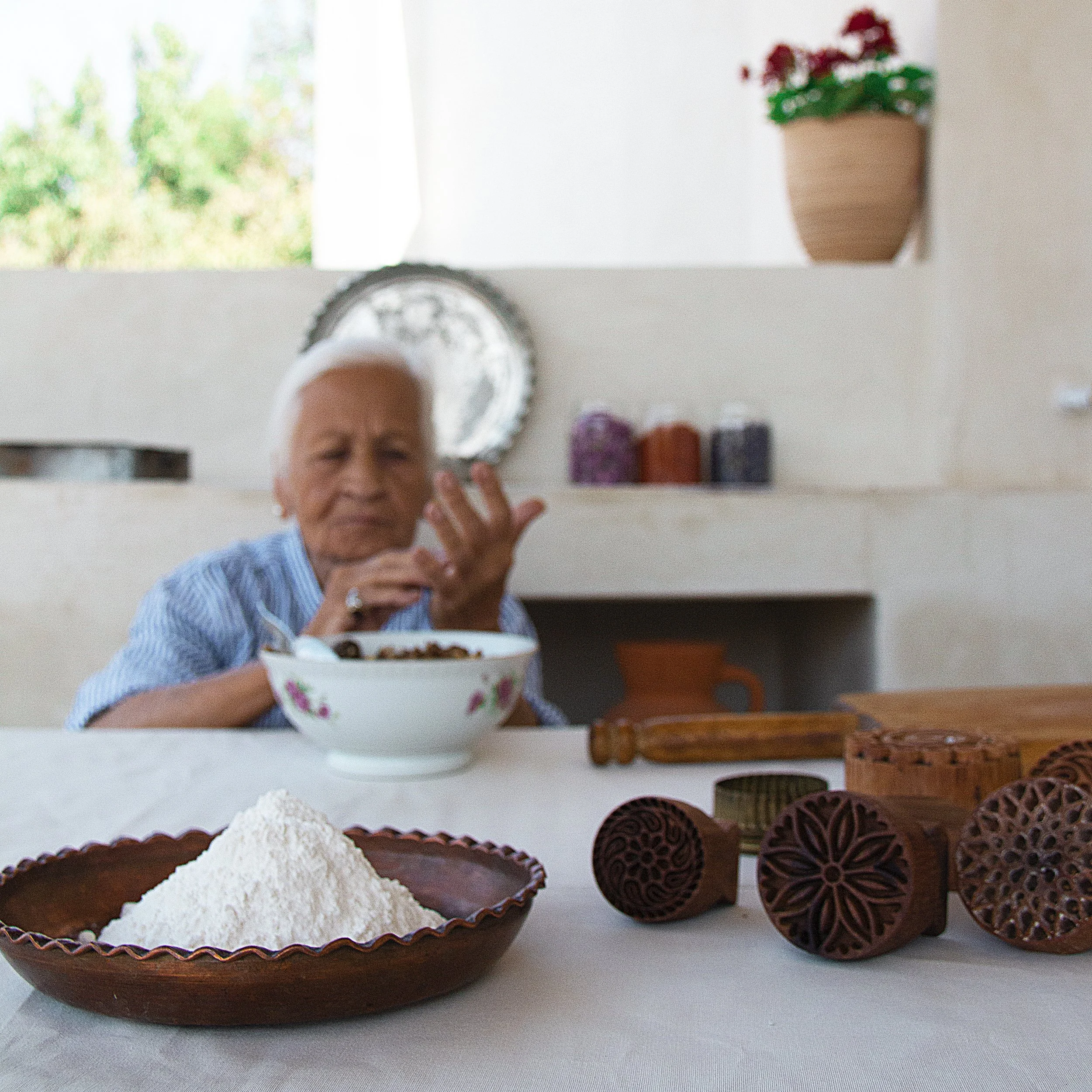 my mom sitting at a table with wooden stamps and a bowl of flour or powder, in a bright room with shelves and a window.