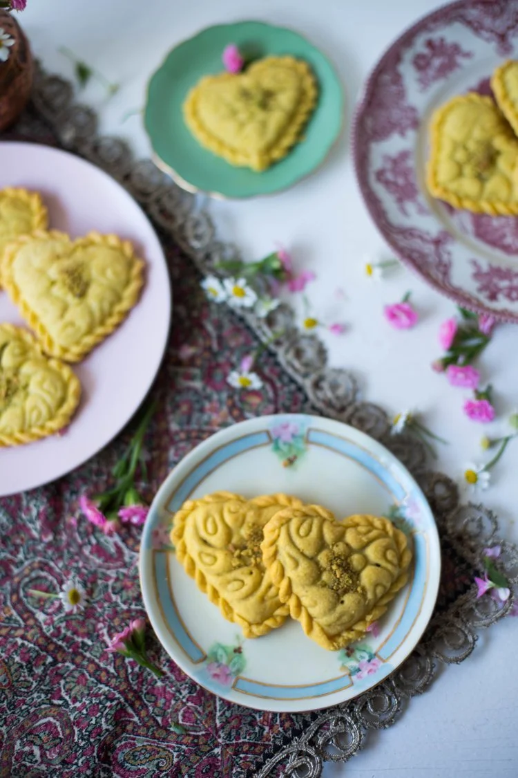 Heart-shaped cookies with decorative patterns on plates surrounded by small pink and white flowers on a decorative tablecloth.