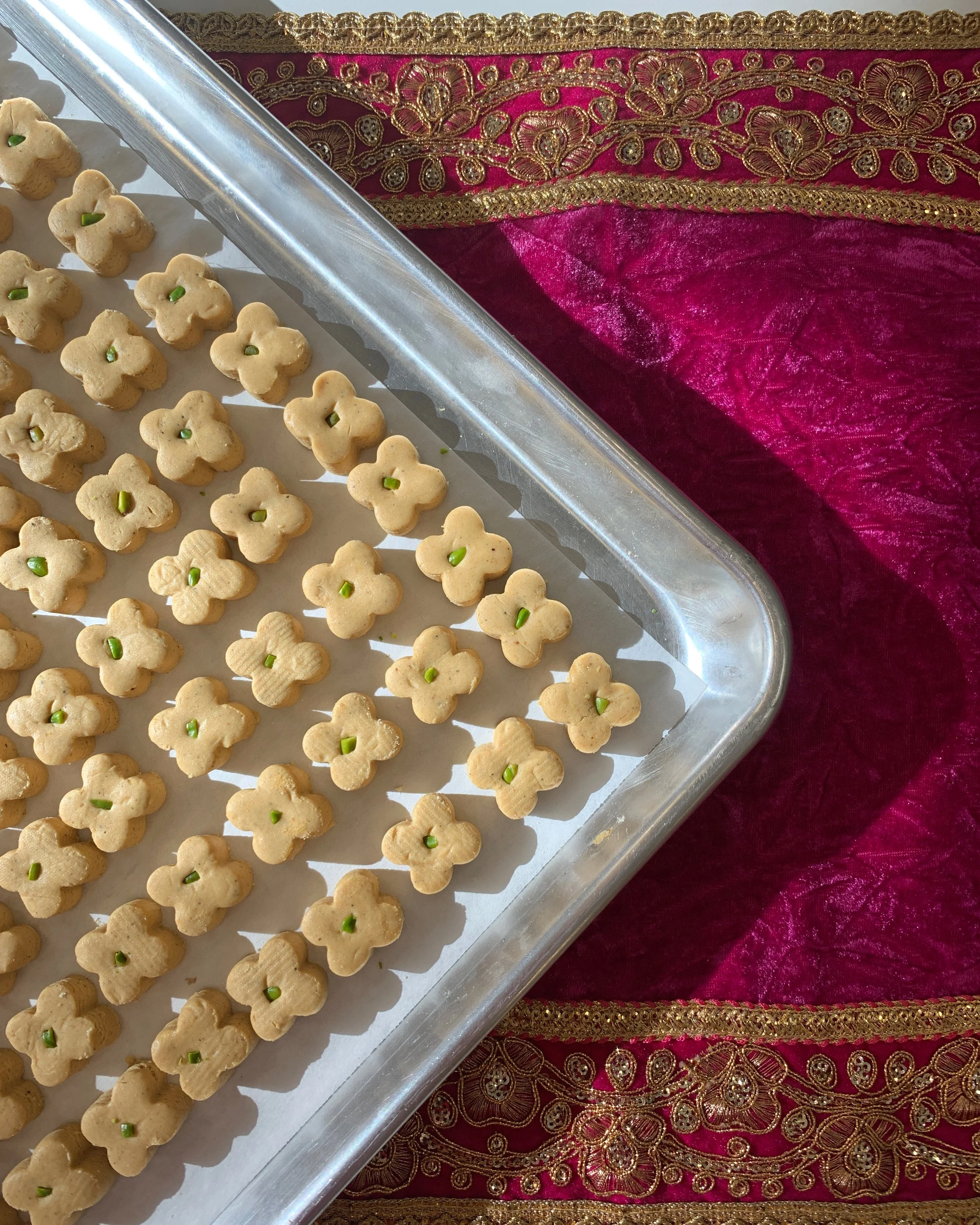 A tray of flower-shaped cookies with a small green piece in the center, placed on a white sheet of paper, next to a pink and gold embroidered fabric.