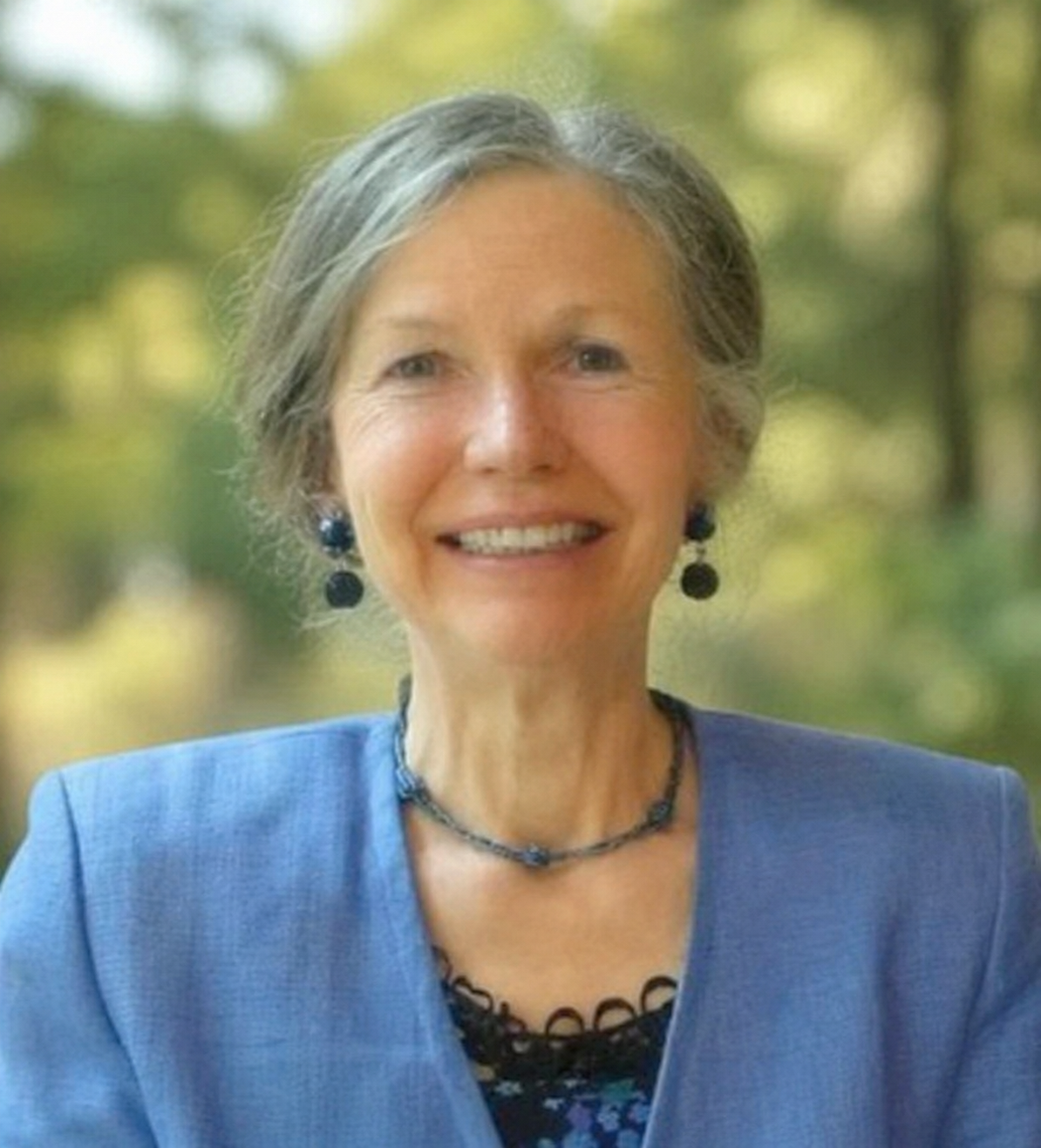 Portrait of an elderly woman with short gray hair, wearing a blue blazer, black necklace, and black earrings, smiling outdoors with blurred green trees in the background.