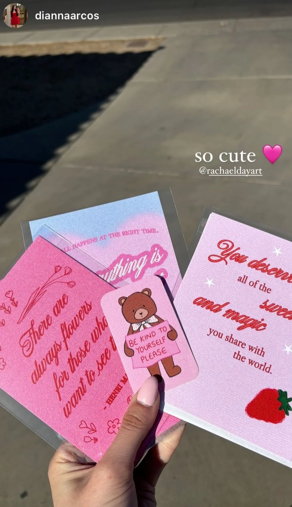 Hand holding three cards with inspirational and cute messages, including a bear sticker holding a sign that says 'Be kind to yourself please' and a strawberry illustration, against a concrete sidewalk background.
