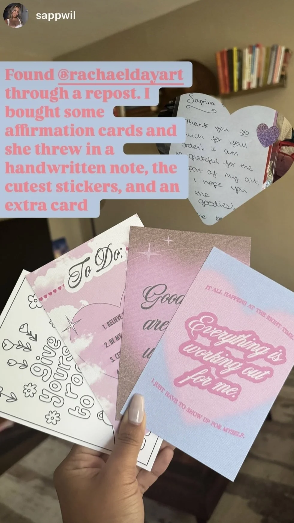 A hand holding several affirmation cards with uplifting messages. The cards are pink, purple, and white with decorative fonts and illustrations. In the background, there is a handwritten note on a heart-shaped card and a bookshelf.