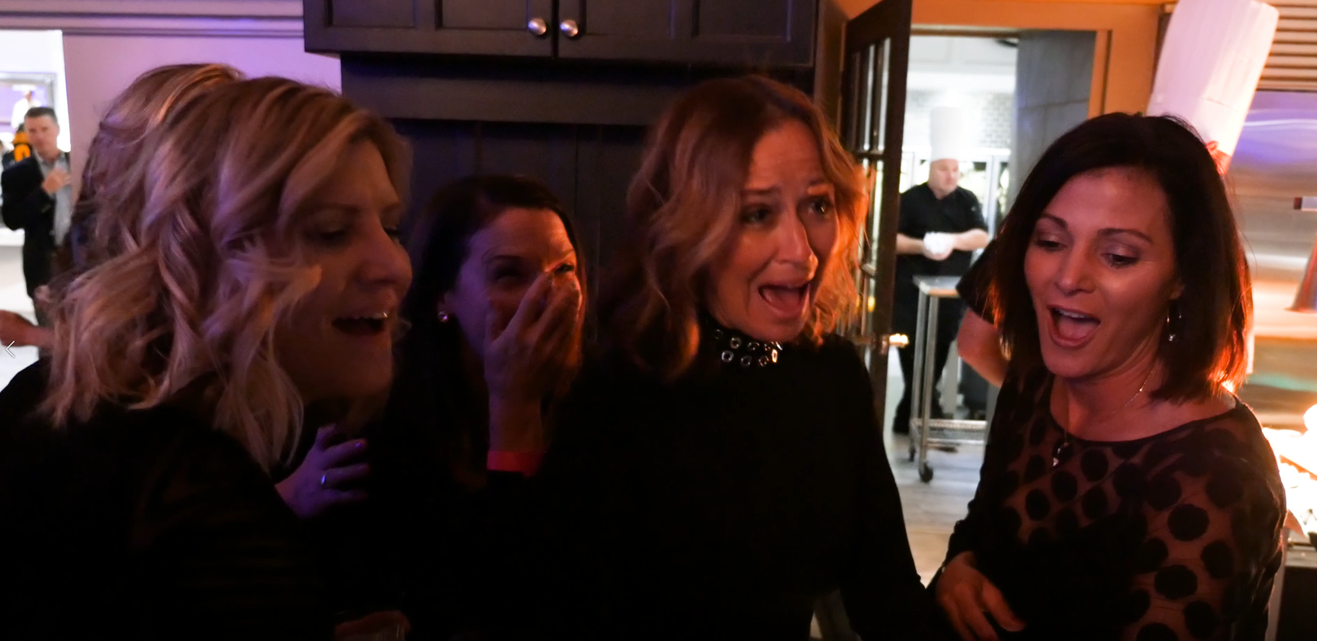 Four women in a dimly lit setting appear surprised and excited, with one woman covering her mouth in shock and the others smiling or reacting to a magician.