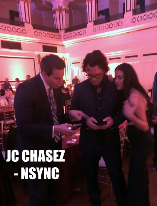 Three people in formal attire looking at their phones in a decorated event space with tables and chairs, pink lighting, and multiple guests seated in the background.