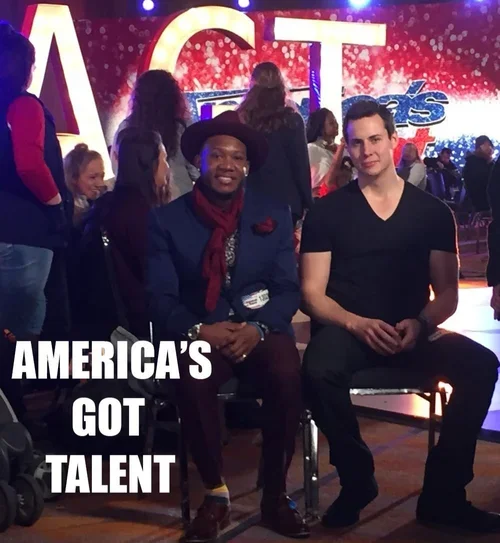 Two men sitting on chairs in front of a stage with a large sign that reads "AMERICA'S GOT TALENT." One man is smiling and wearing a hat and scarf, the other is looking at the camera and wearing a black shirt. Several people are standing and sitting b