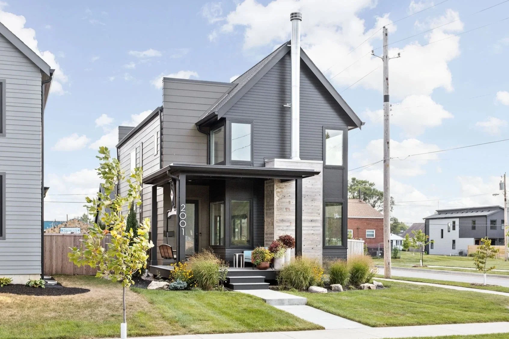 Modern two-story house with gray and black exterior, front yard with lap lawn, small trees, flowers, and a porch with potted plants and outdoor furniture. Street and neighboring houses are in the background.