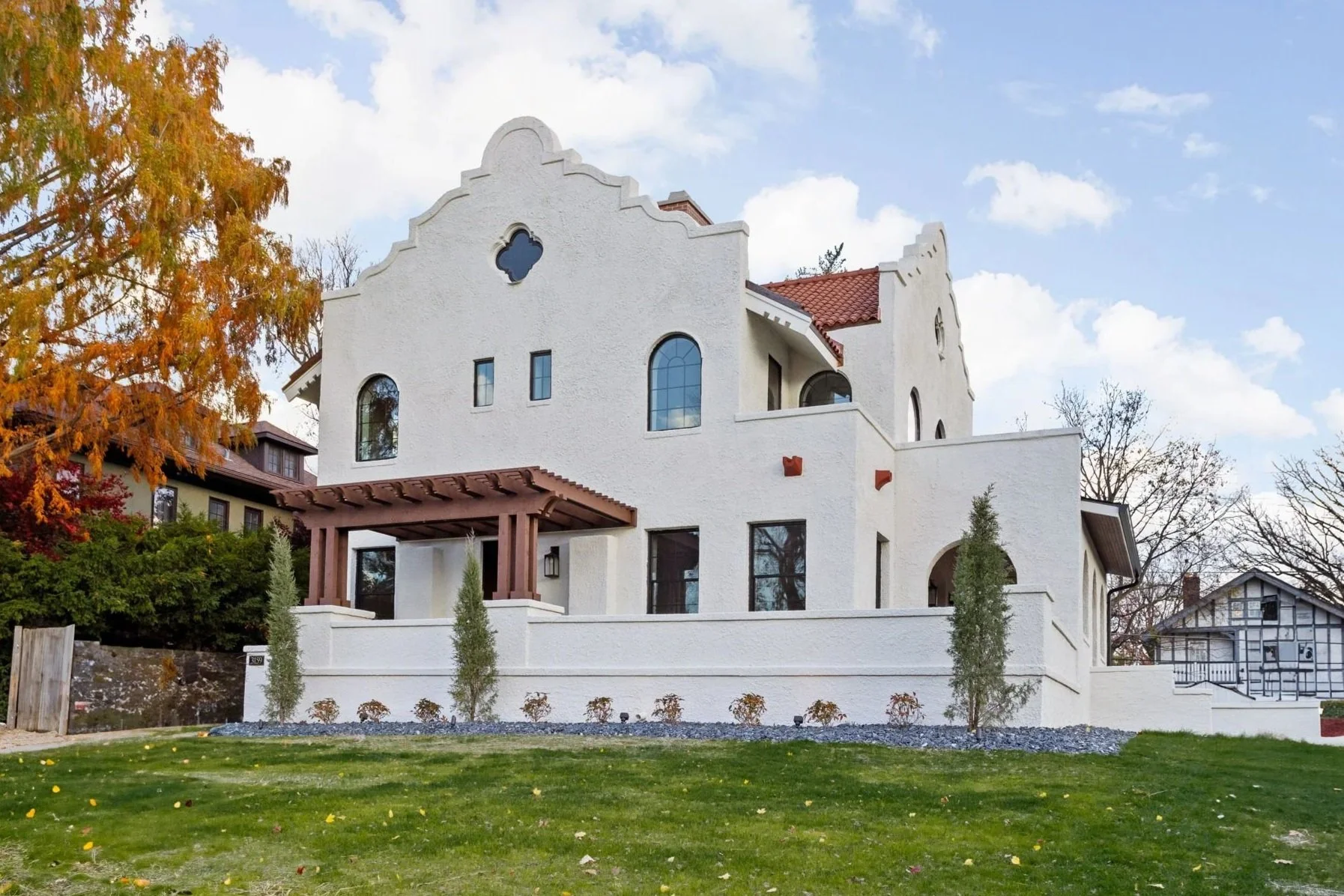 A large white stucco house with a red tile roof, multiple arched and rectangular windows, and a small balcony. A front lawn has scattered leaves and small trees, with a wooden pergola at the entrance. The background shows a blue sky with white clouds and trees with autumn foliage.