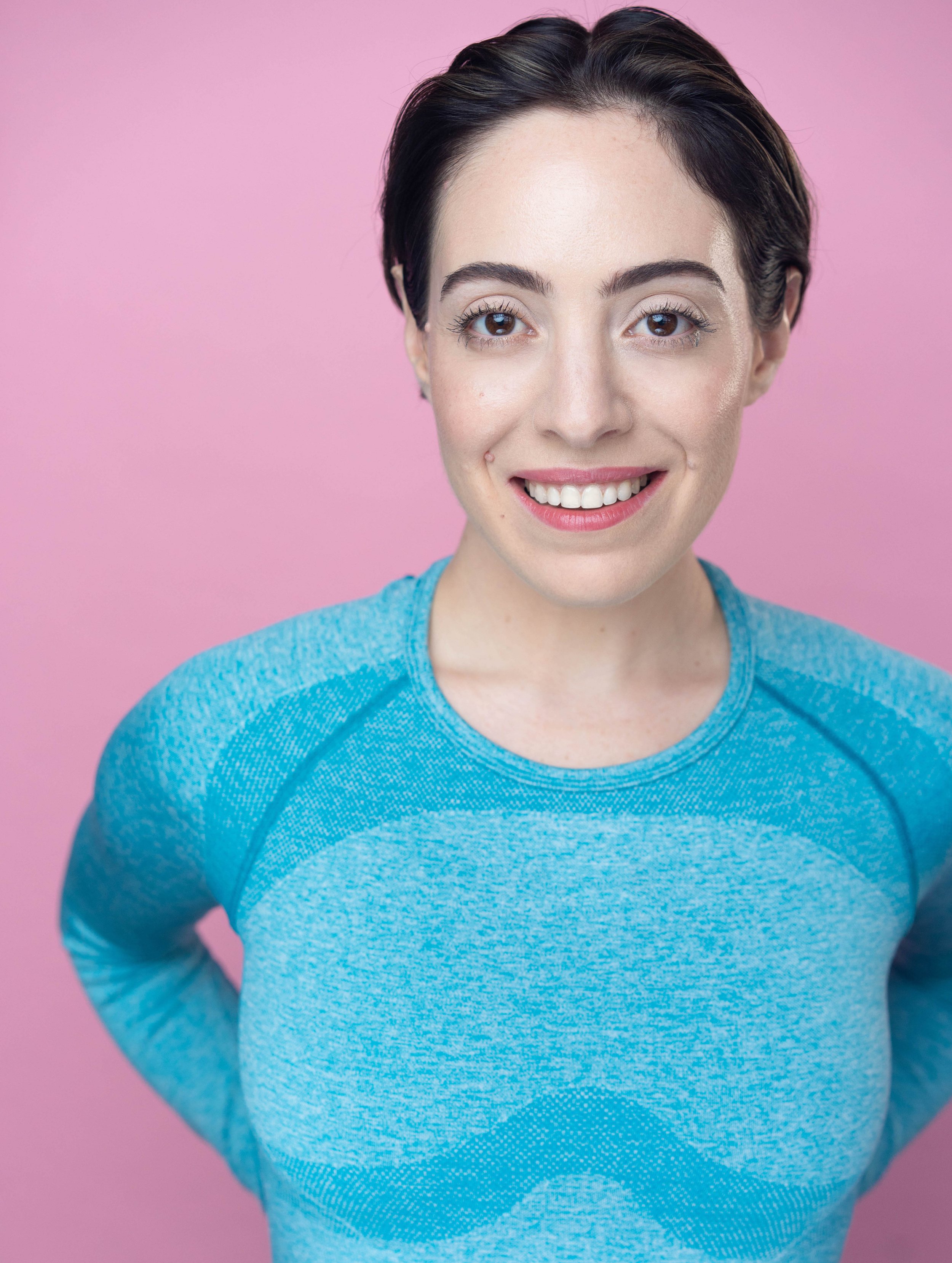  Commercial headshot of actor Yvonne Cone with a pink background, wearing a bright blue, form-fitting, long sleeve workout top. 
